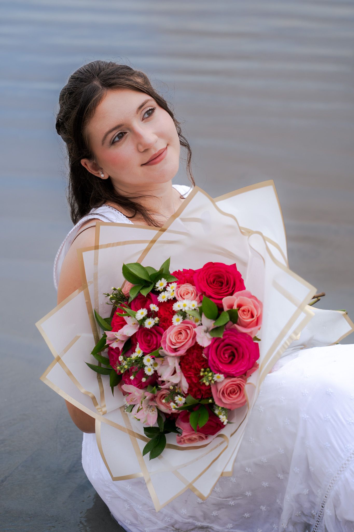 Young woman in white dress sitting by water holding a bouquet of pink and red roses.