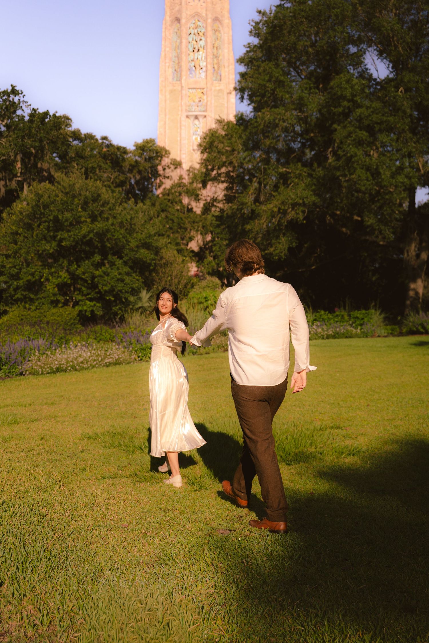 A couple holding hands and walking across a sunny green lawn near a tall historic tower.