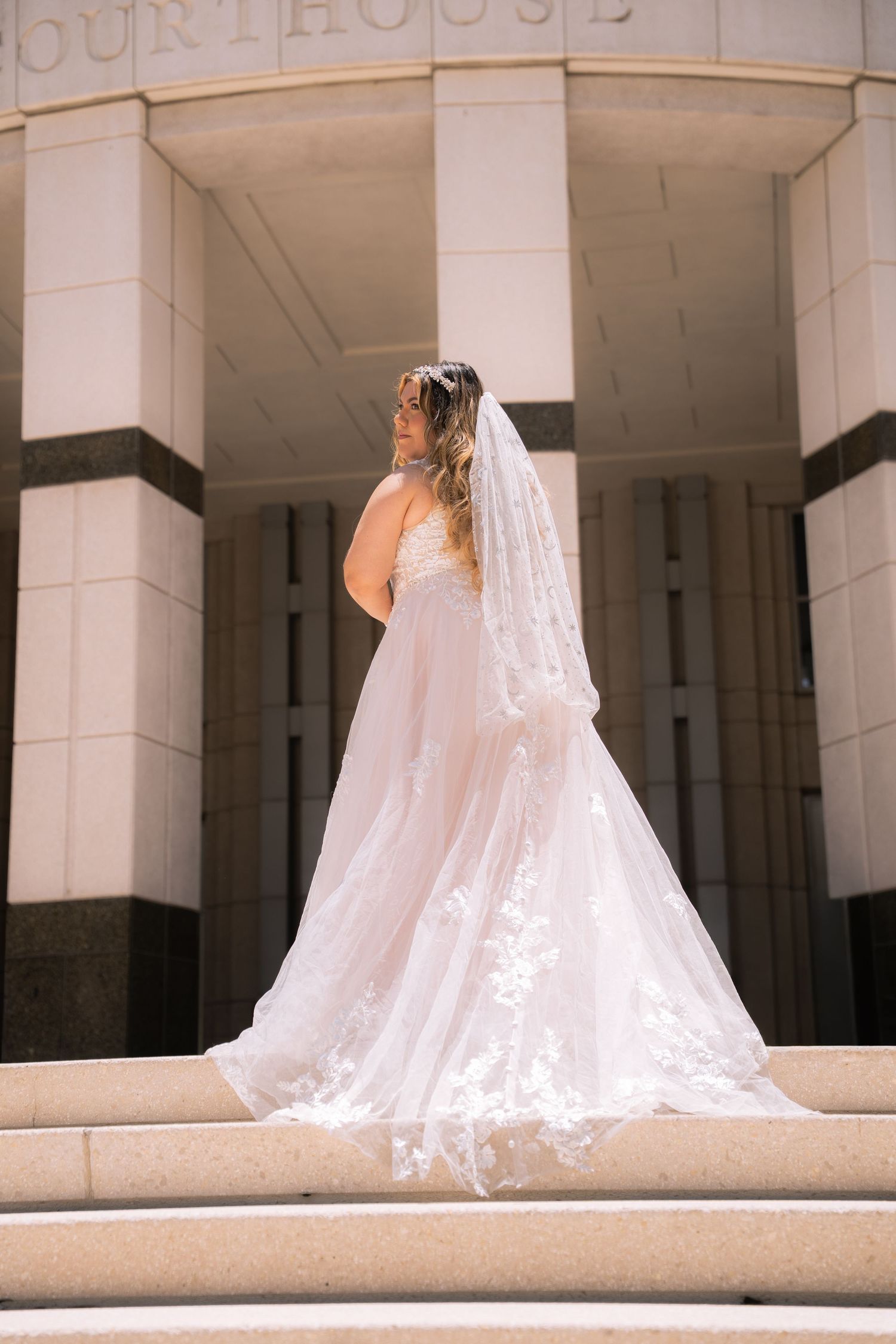 Bride in flowing white lace gown with veil standing on courthouse steps in sunlight.