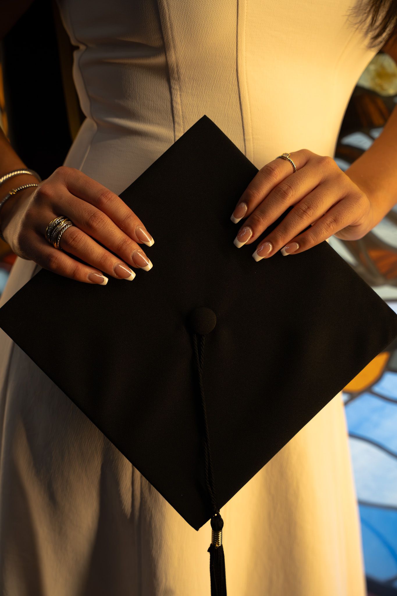 Close-up of graduate hands holding black mortarboard cap, wearing rings and bracelet in white dress.