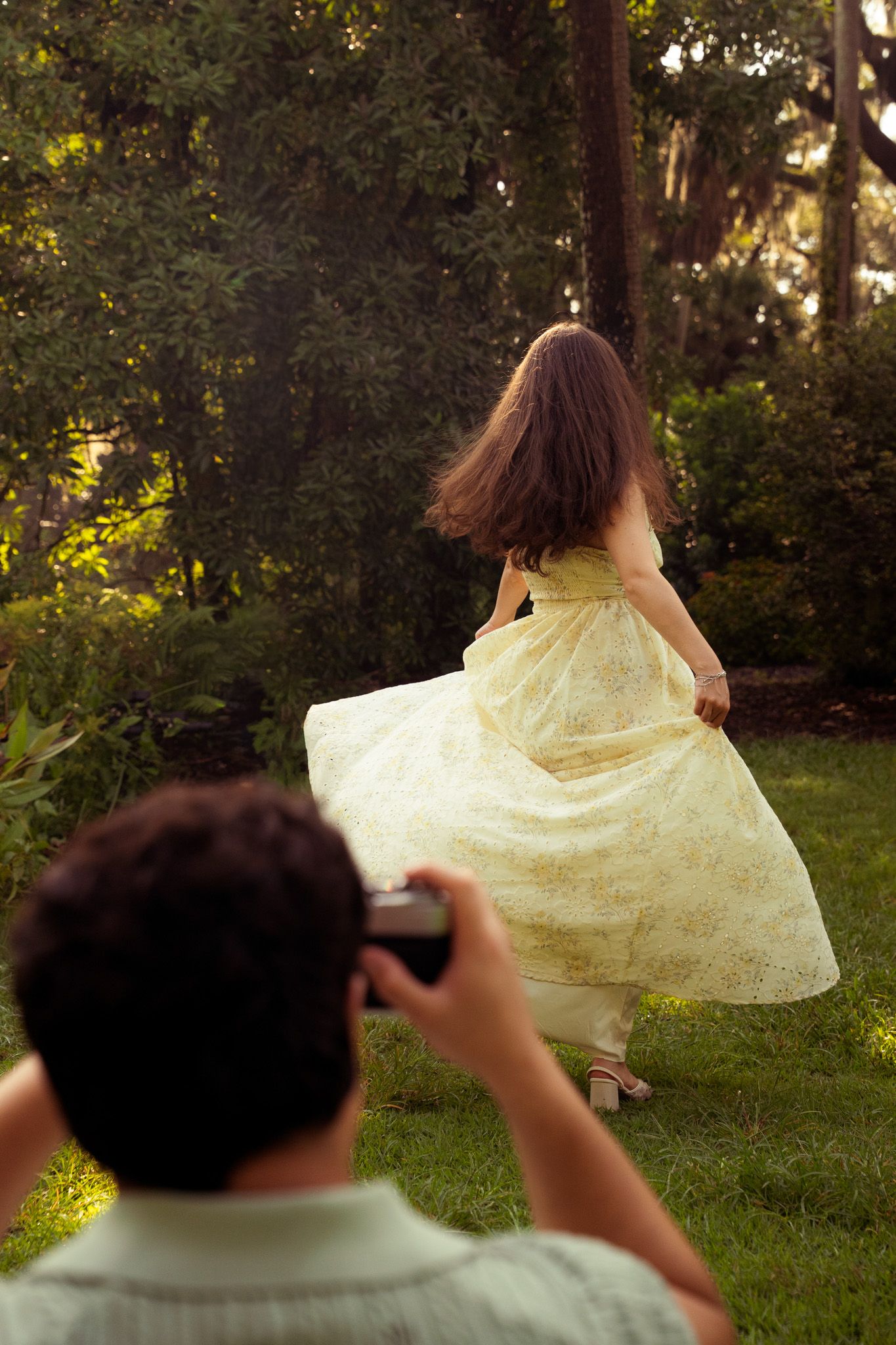 Man photographing woman in a flowing yellow floral dress twirling in a lush green garden.