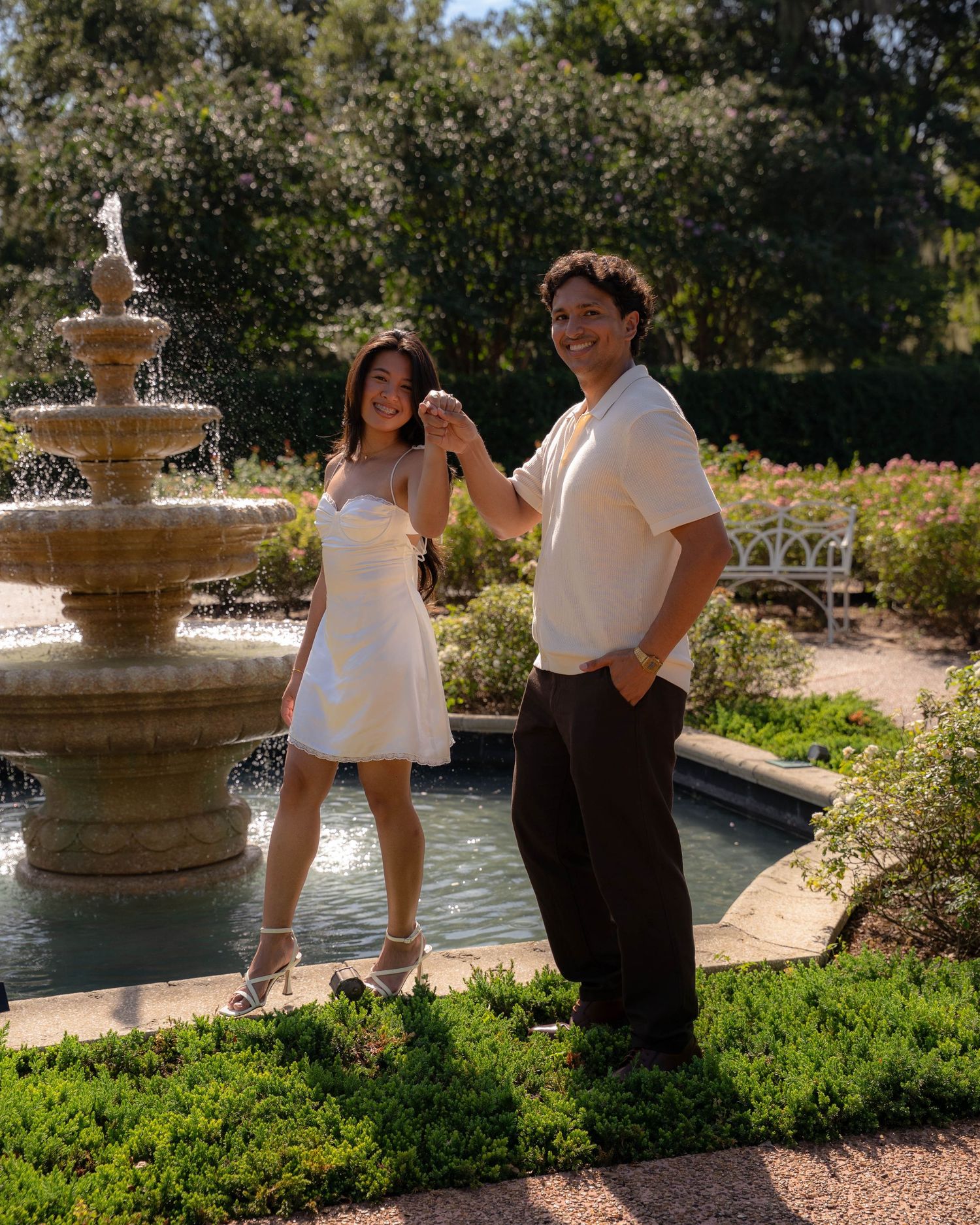 Happy couple dancing near ornate stone fountain in a beautiful garden, woman in white mini dress.