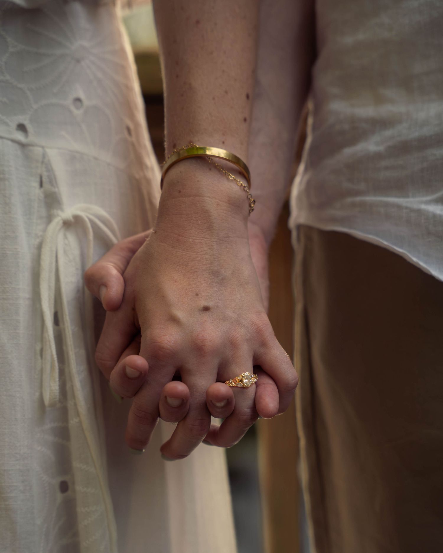 Close-up of couple holding hands, woman wearing gold bangle bracelet and floral ring, both in white.
