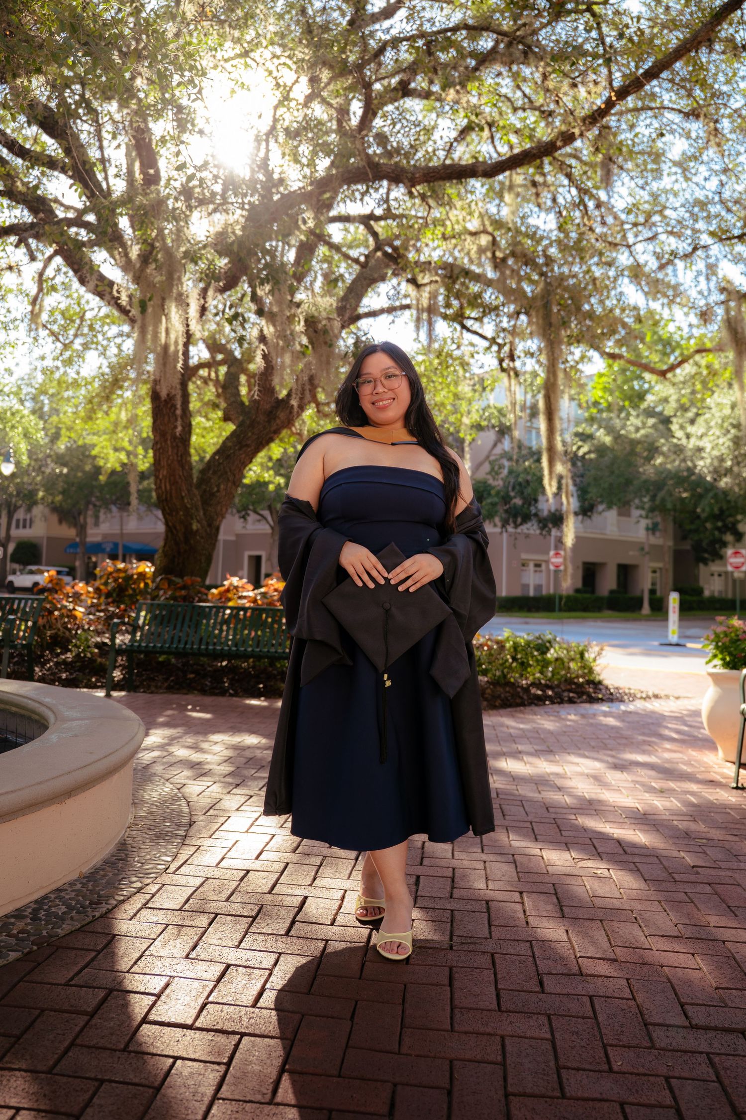 Graduate in navy strapless dress holds black academic gown open under sunlit moss-draped oak trees.