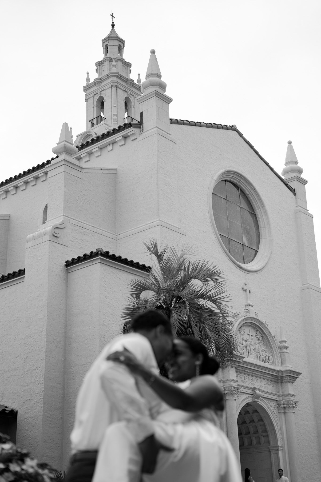 Black and white photo of couple embracing in front of a historic church facade.