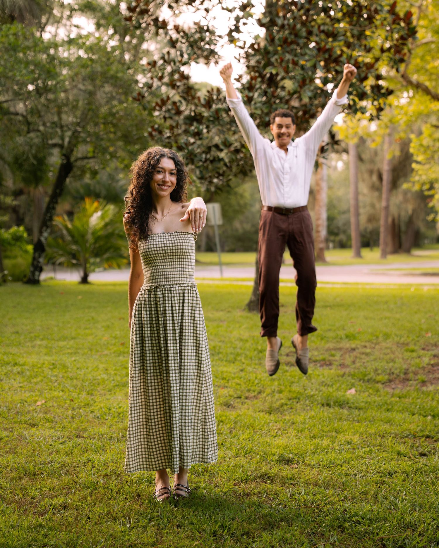 Happy couple in park, woman in green gingham dress showing ring, man jumping joyfully behind her.