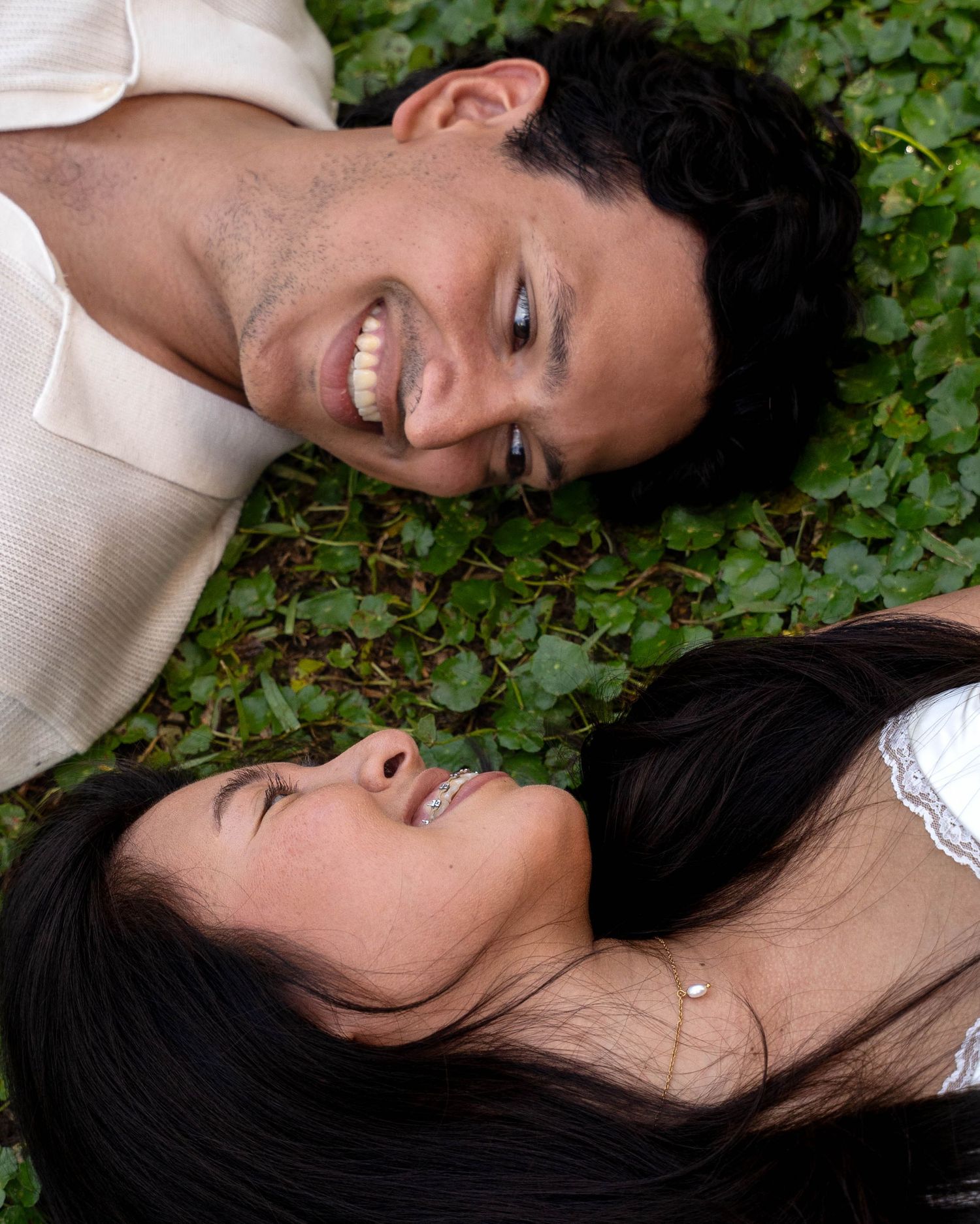 Smiling couple lying on green ivy ground looking at each other from opposite directions outdoors.