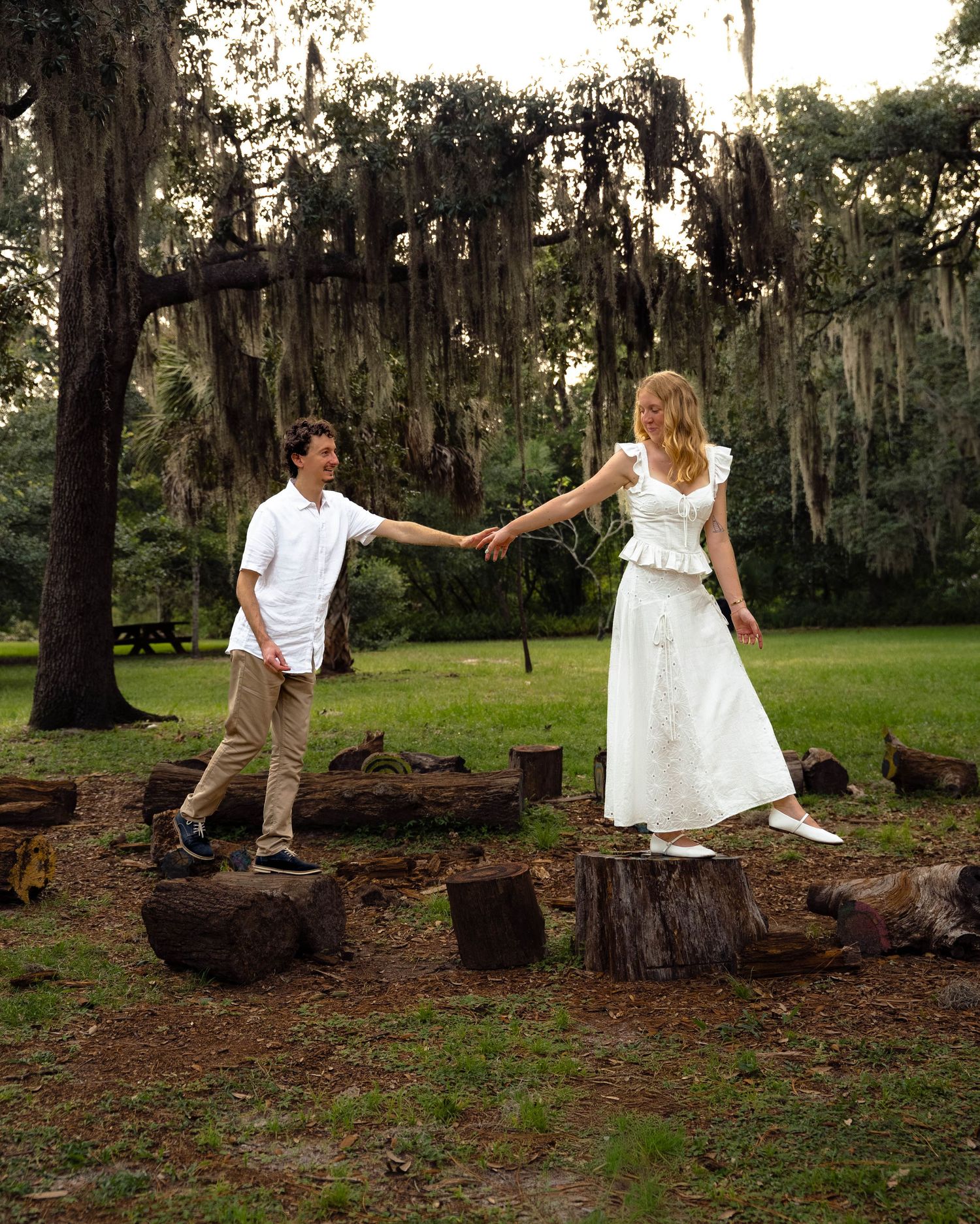 Couple holding hands while balancing on tree stumps in a mossy park at sunset.
