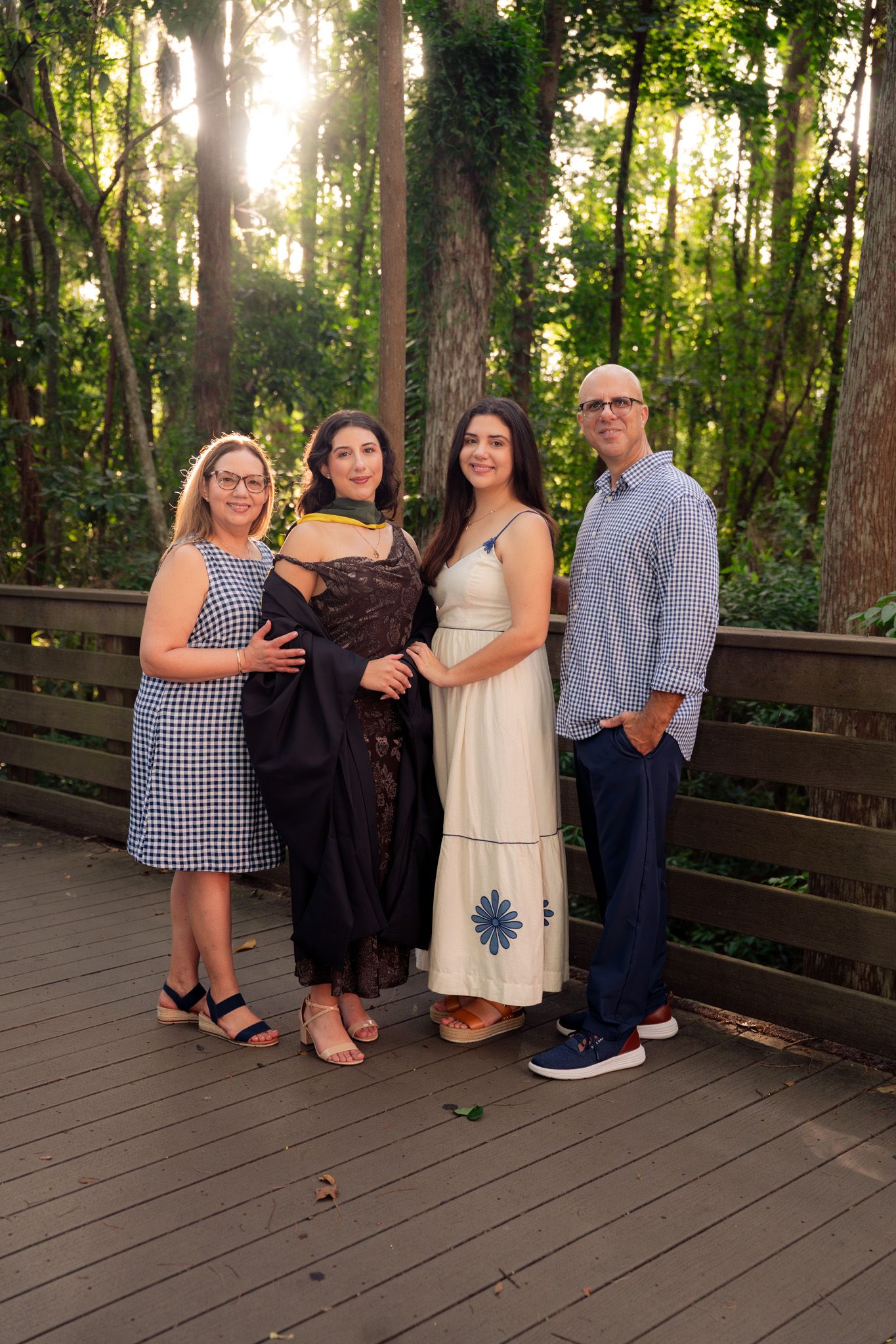 Family of four poses on wooden bridge surrounded by sunlit forest greenery.