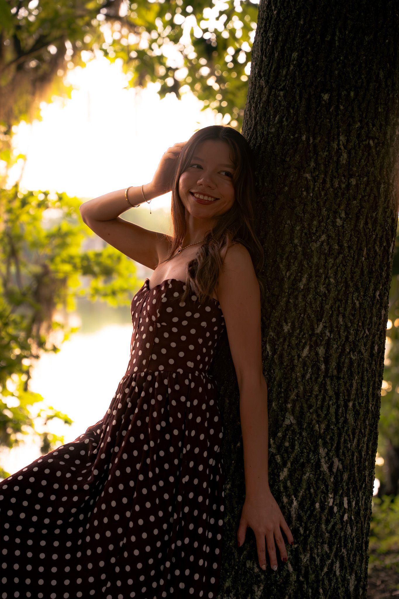 Joyful woman in burgundy polka dot maxi dress leaning against tree trunk in golden hour park light.