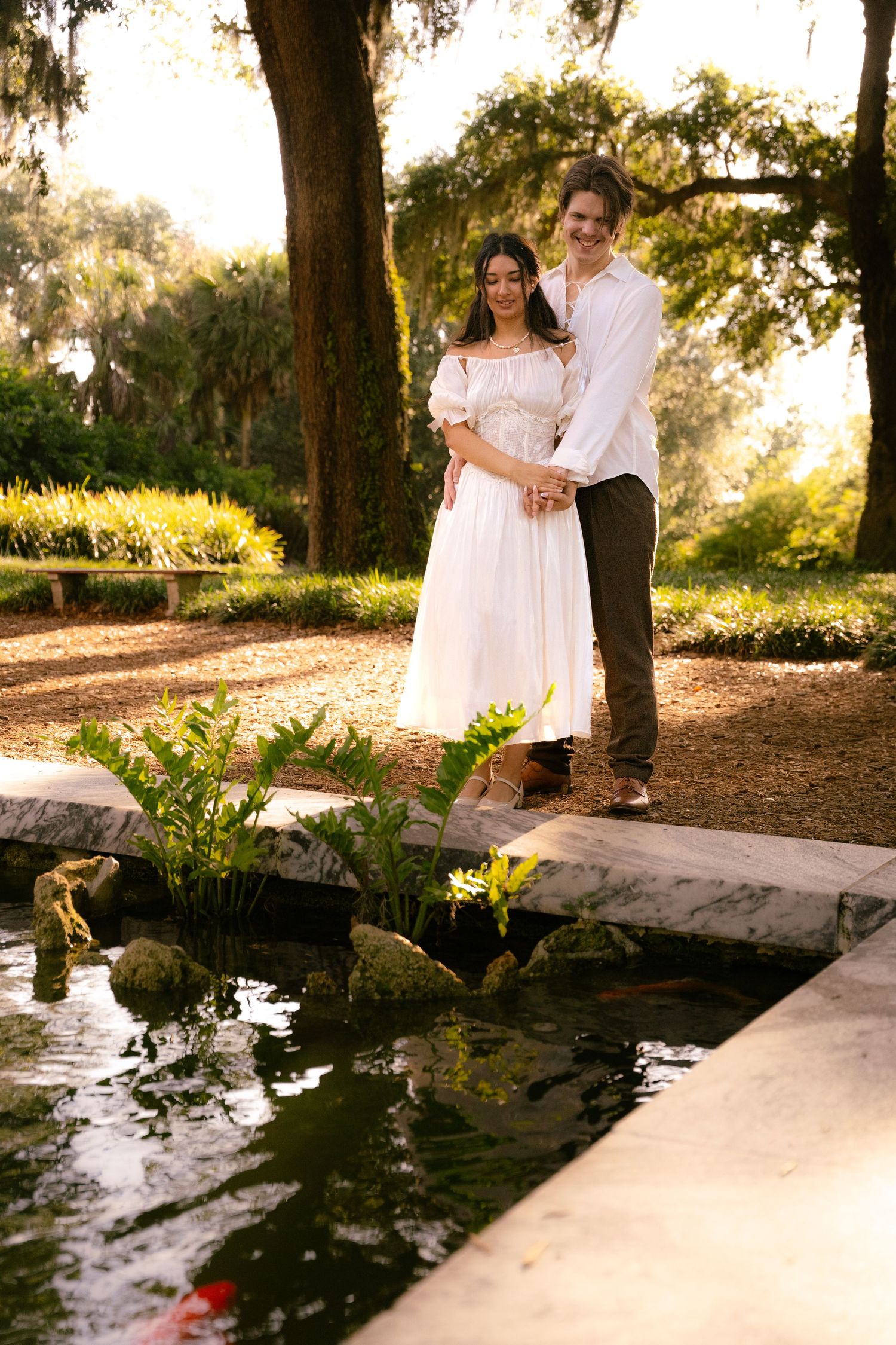 Couple standing together by a koi pond in a beautiful sunlit garden setting.
