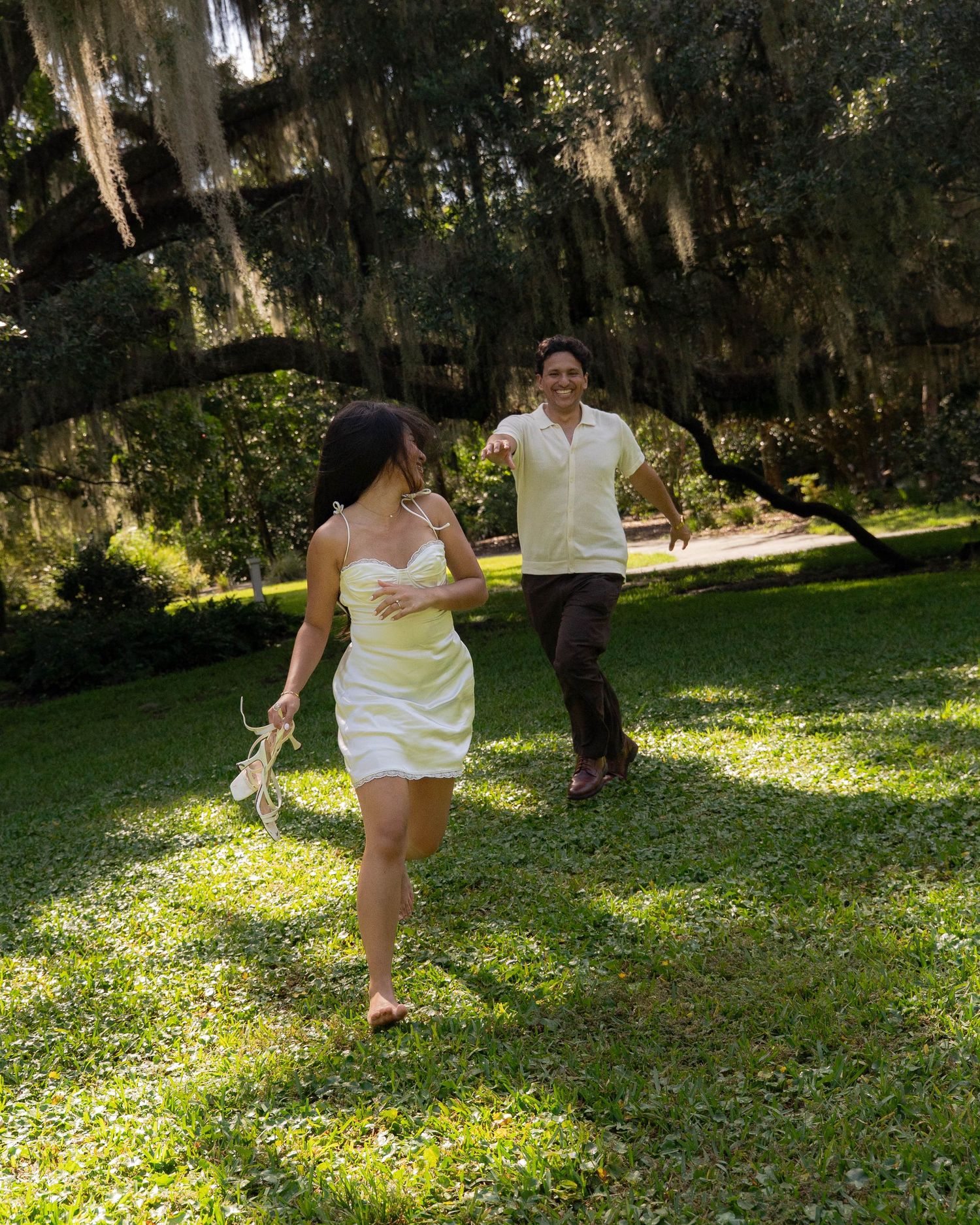 Couple running barefoot through a sunlit park with Spanish moss-draped trees.