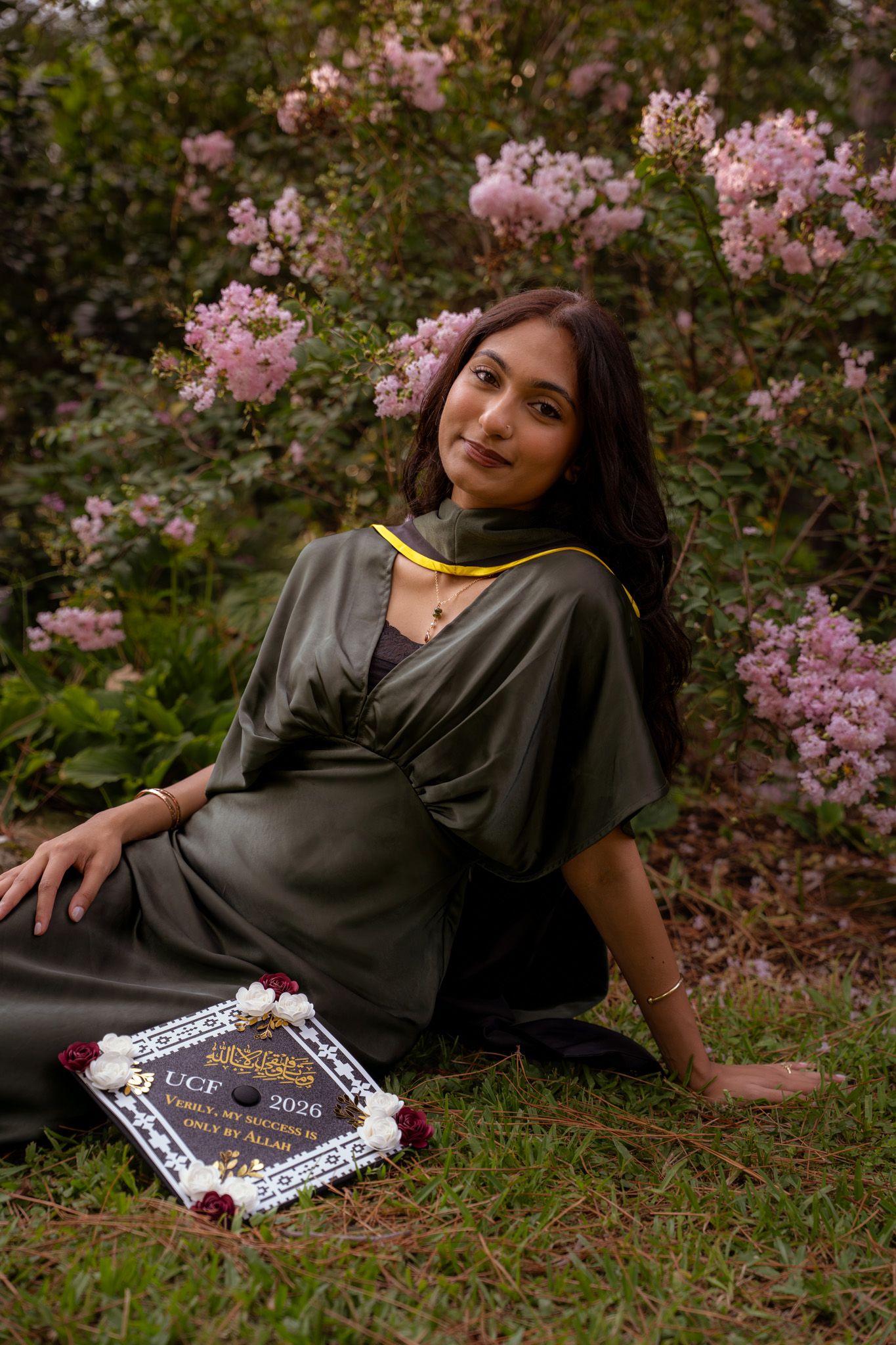 Young woman in olive graduation gown sits among pink blooming flowers holding a UCF 2026 decorated cap.