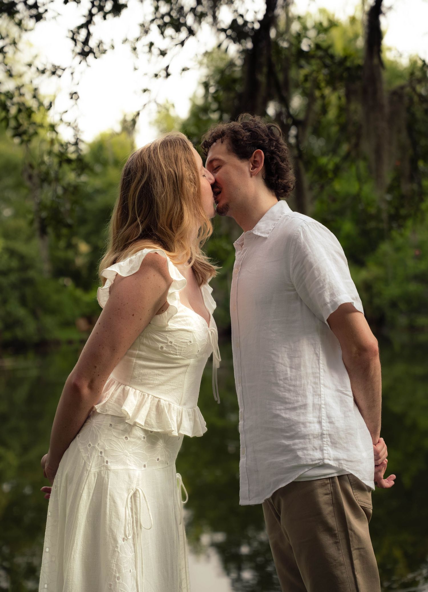 Couple sharing a romantic kiss near a reflective pond surrounded by lush green trees.