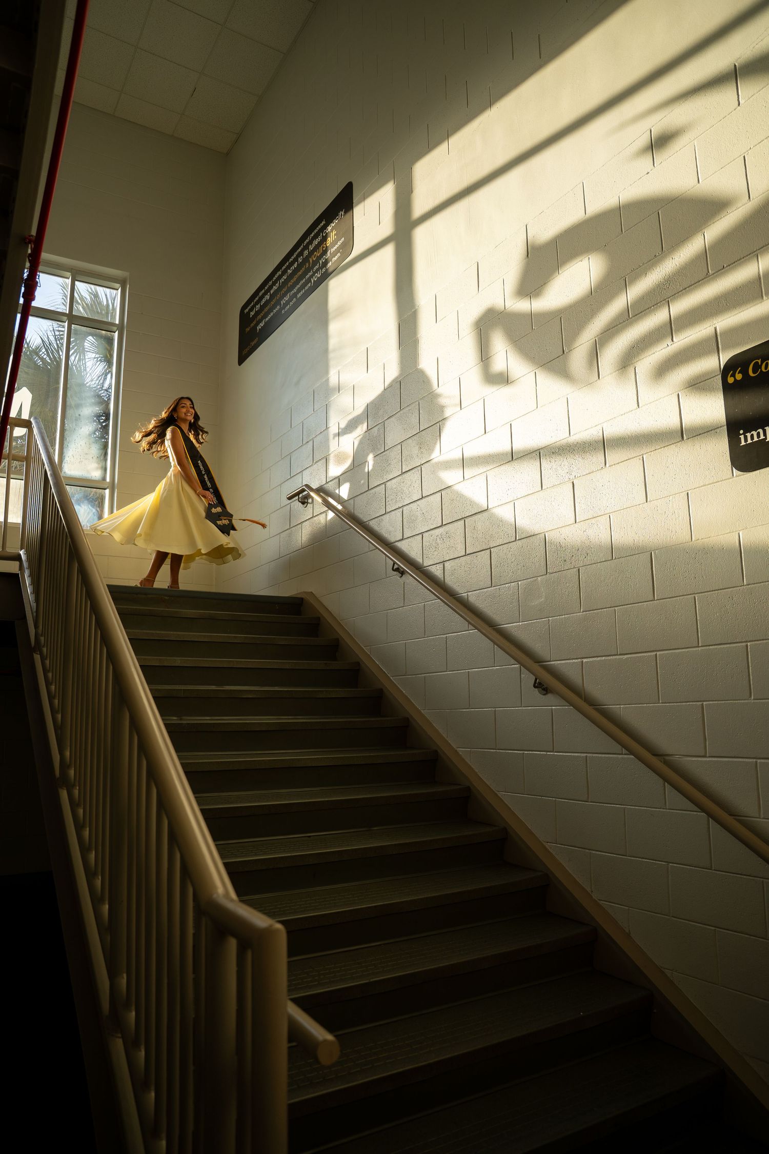 Woman in yellow dress descends sunlit staircase casting dramatic shadow on white brick wall.