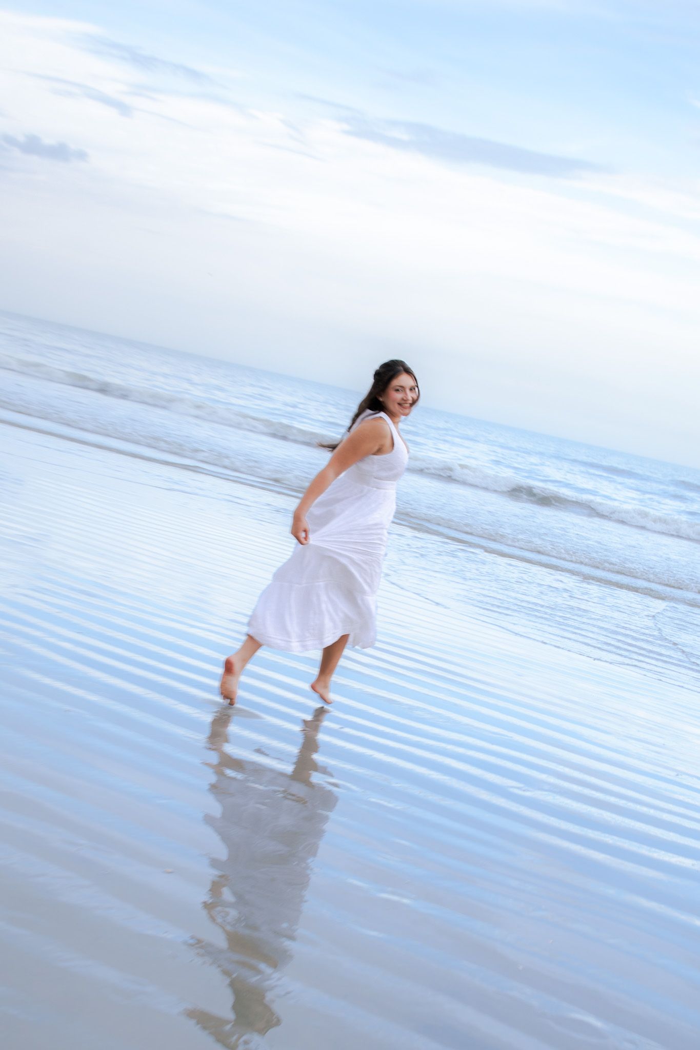Woman in flowing white dress running barefoot along a reflective wet beach at dusk.