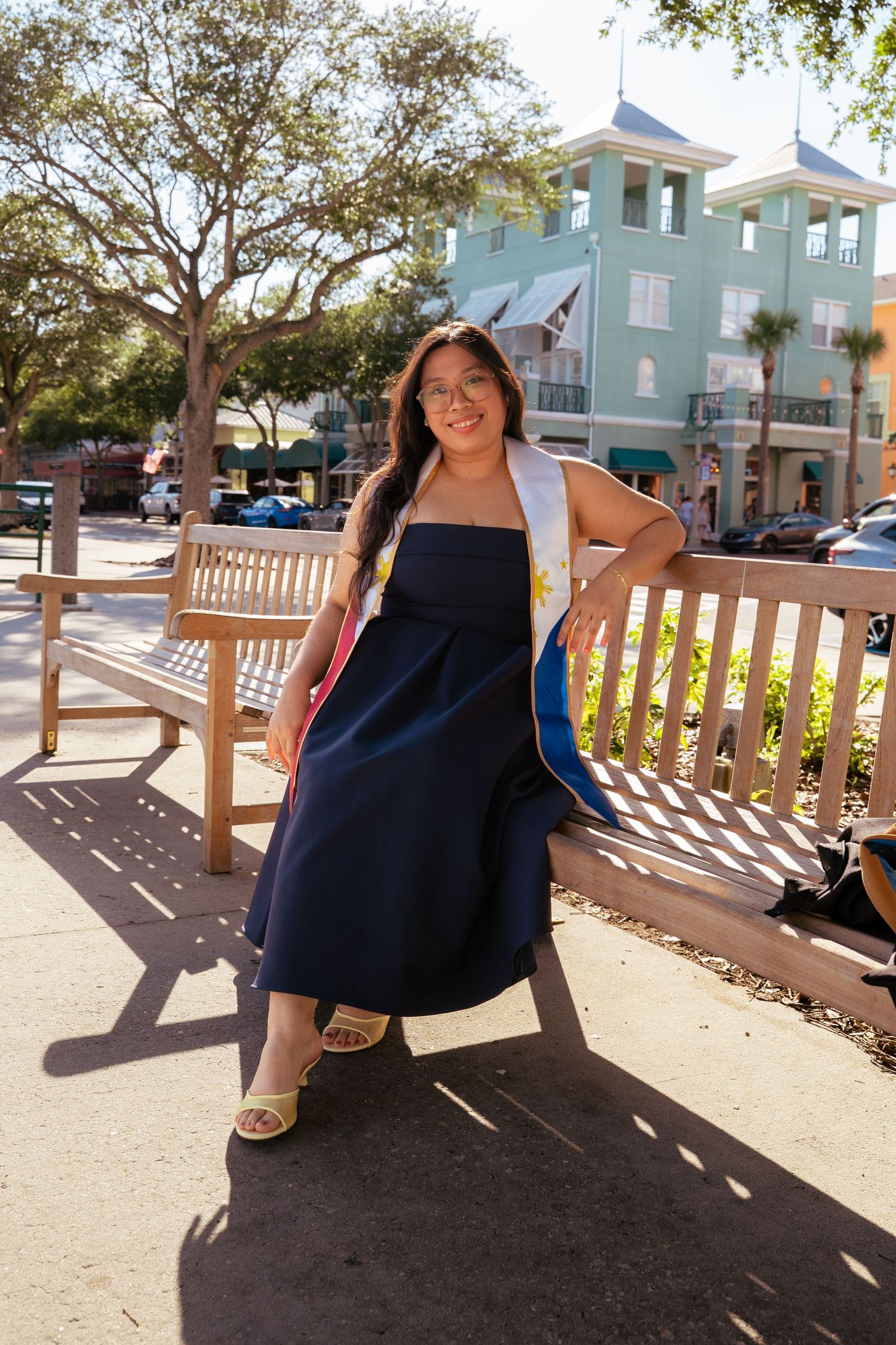 Smiling graduate in navy dress with colorful hood leans against wooden bench in sunny outdoor plaza.