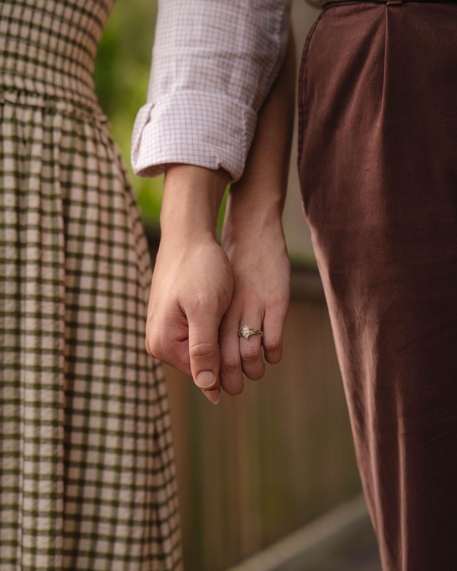 Close-up of couple holding hands, woman wearing diamond engagement ring, dressed in checkered skirt and brown velvet trousers.