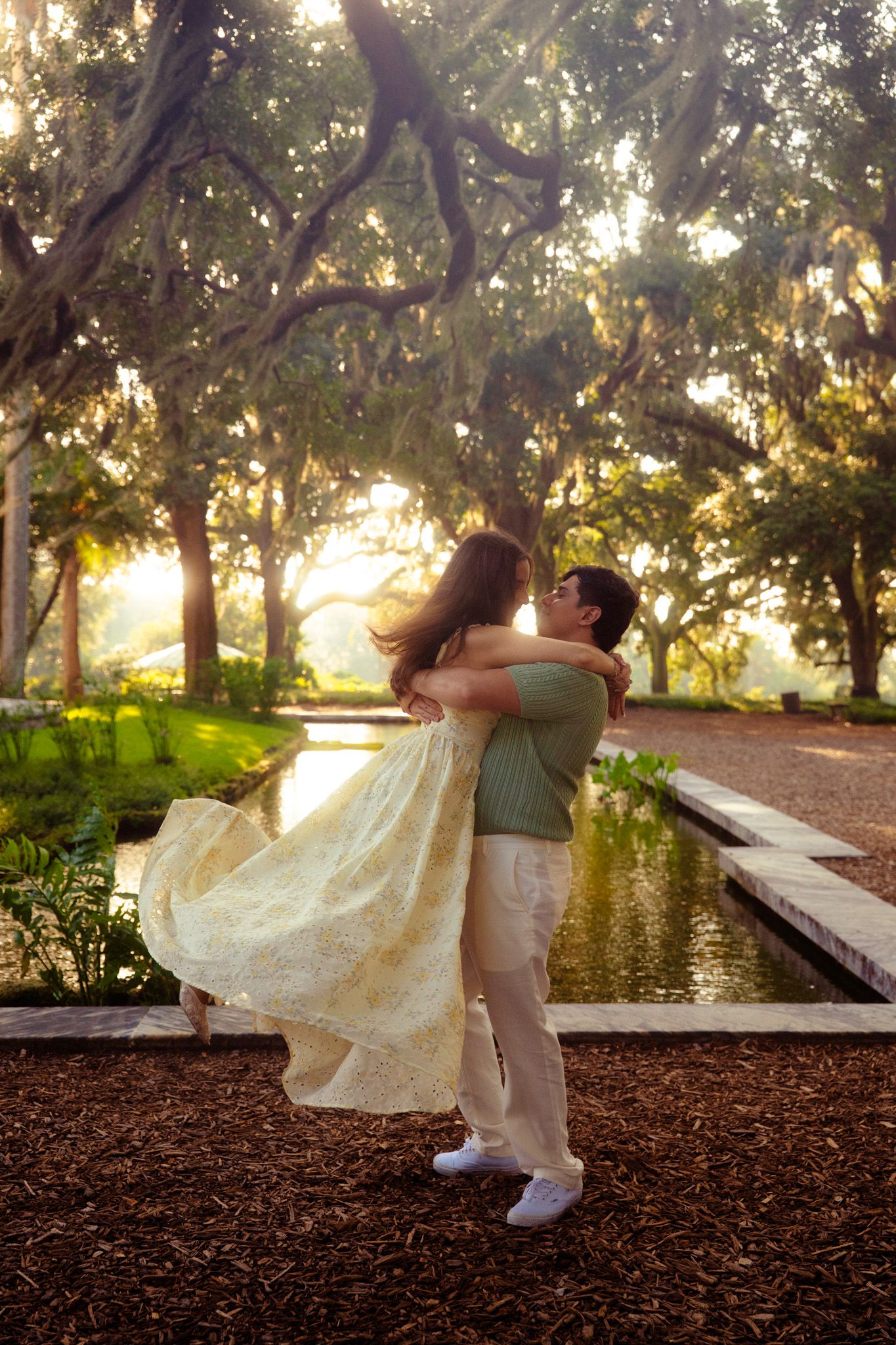 Man lifting woman in yellow dress, spinning her near a pond under oak trees at sunset.