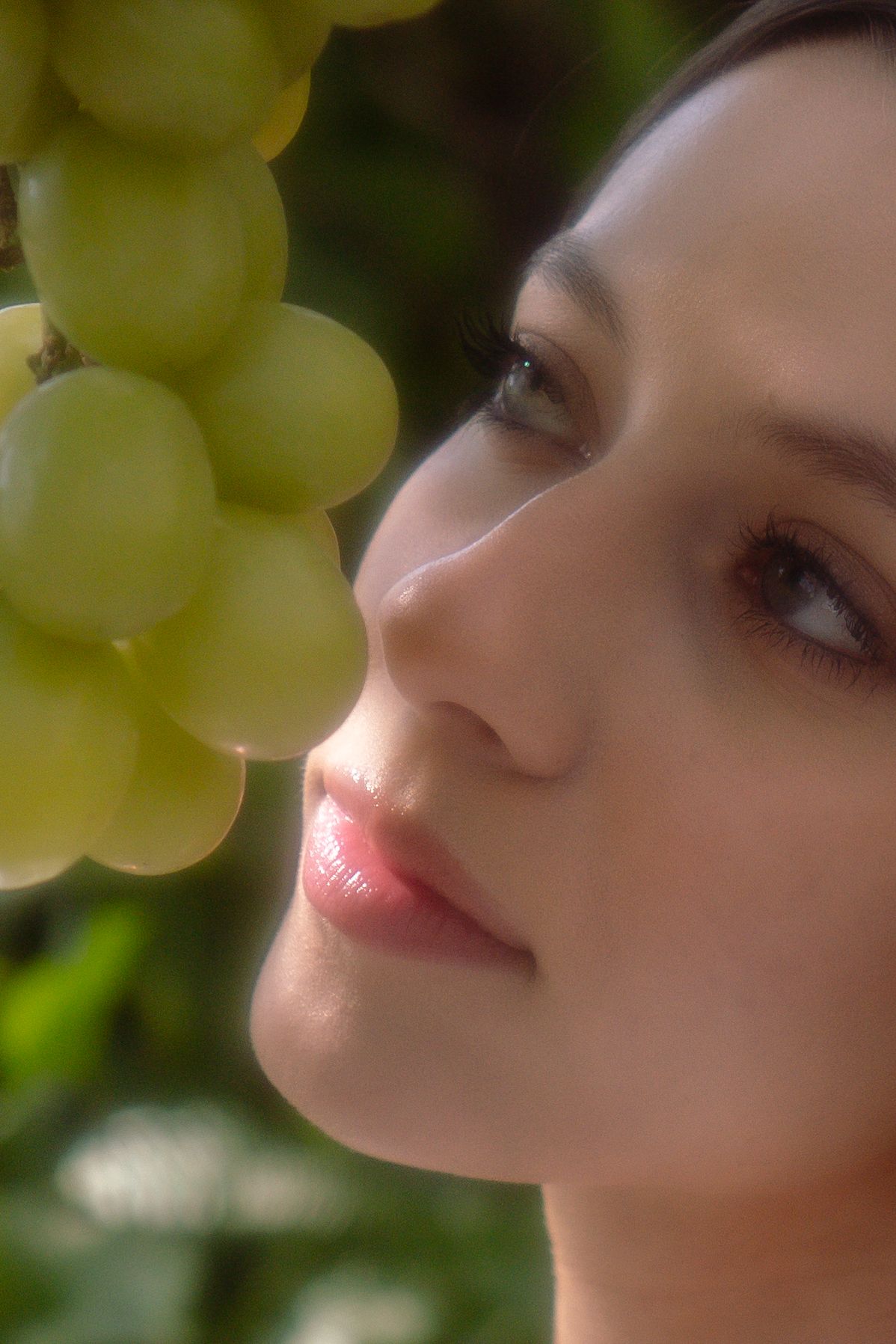 A close-up profile of a young woman with pink lips gazing at a cluster of green grapes.