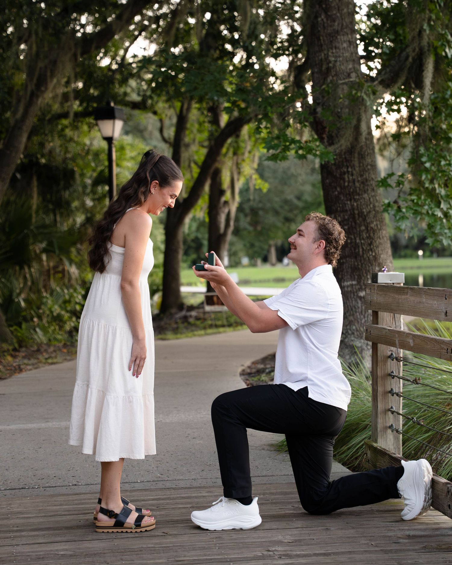 Man kneeling on one knee proposing to woman in white dress on scenic tree-lined path outdoors.
