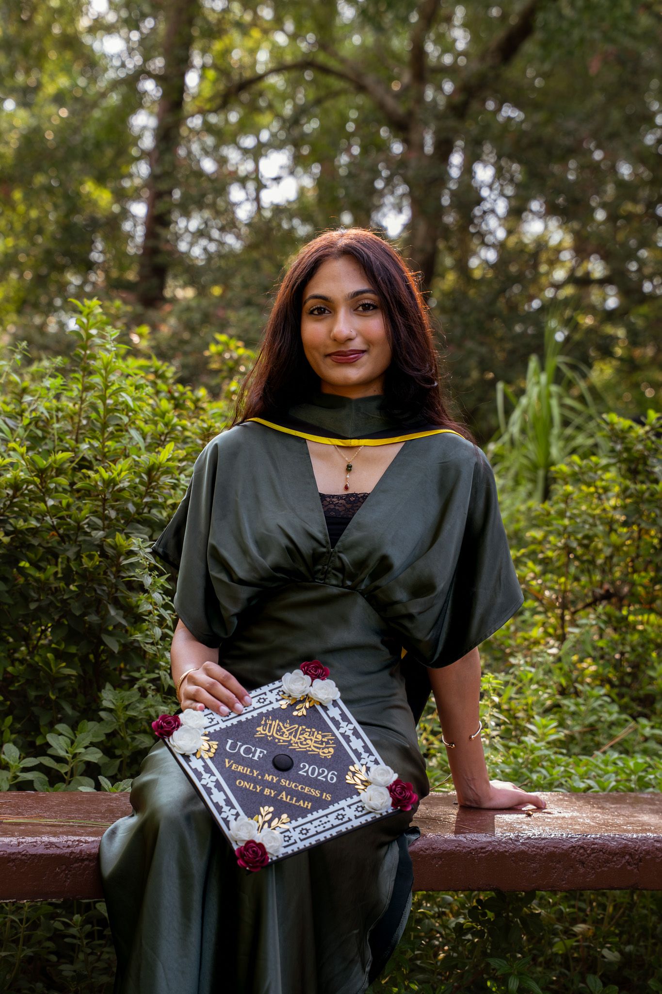 Female UCF 2026 graduate in dark gown sits outdoors holding decorated graduation cap with roses.