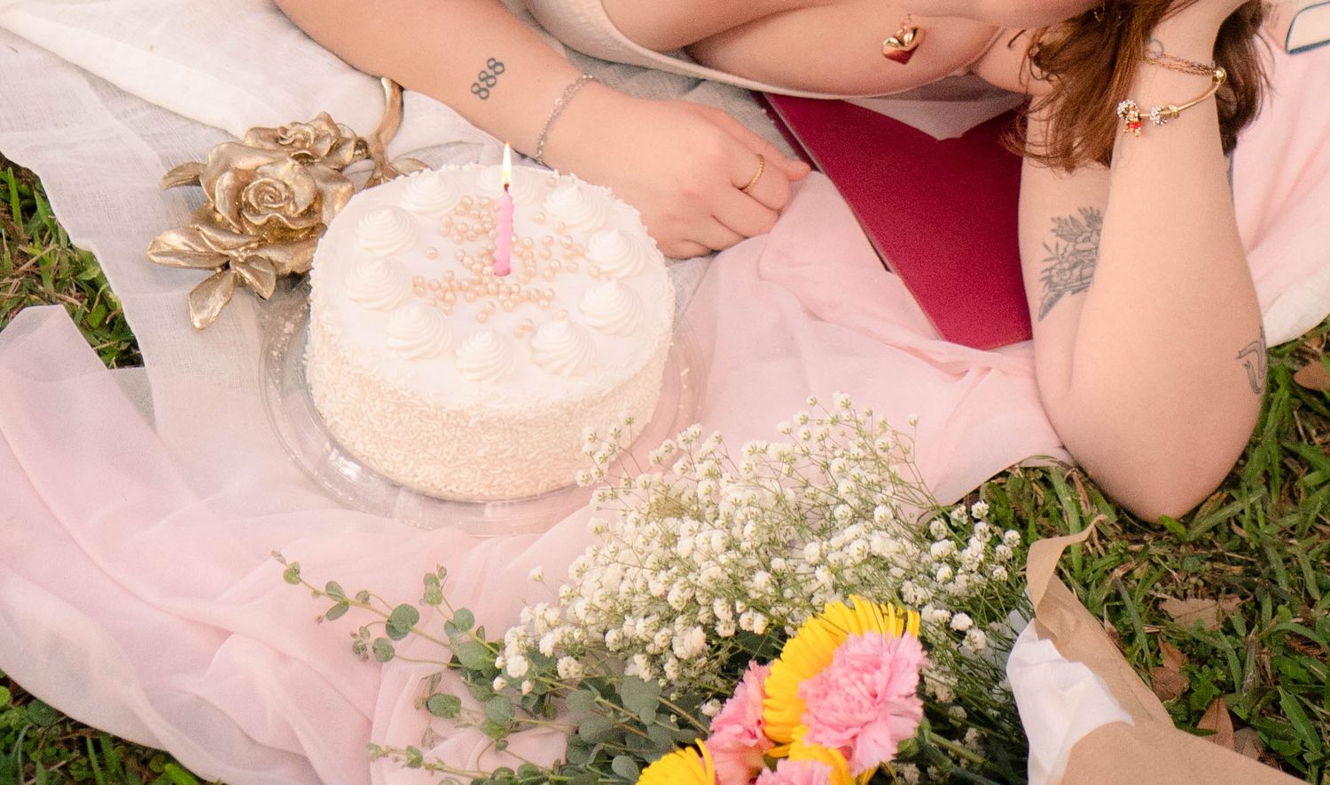 Close-up of a white birthday cake with a candle surrounded by flowers on a pink blanket outdoors.