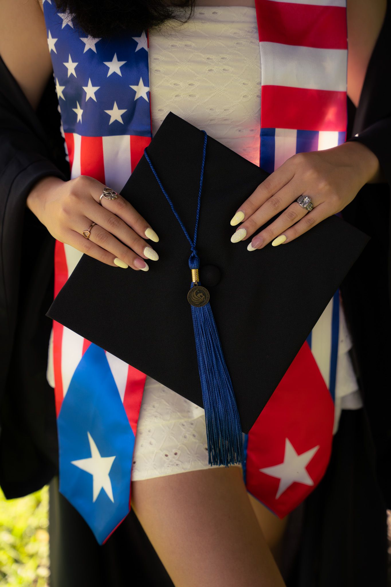 Graduate holds black mortarboard cap wearing American and Puerto Rican flag stoles over white dress.