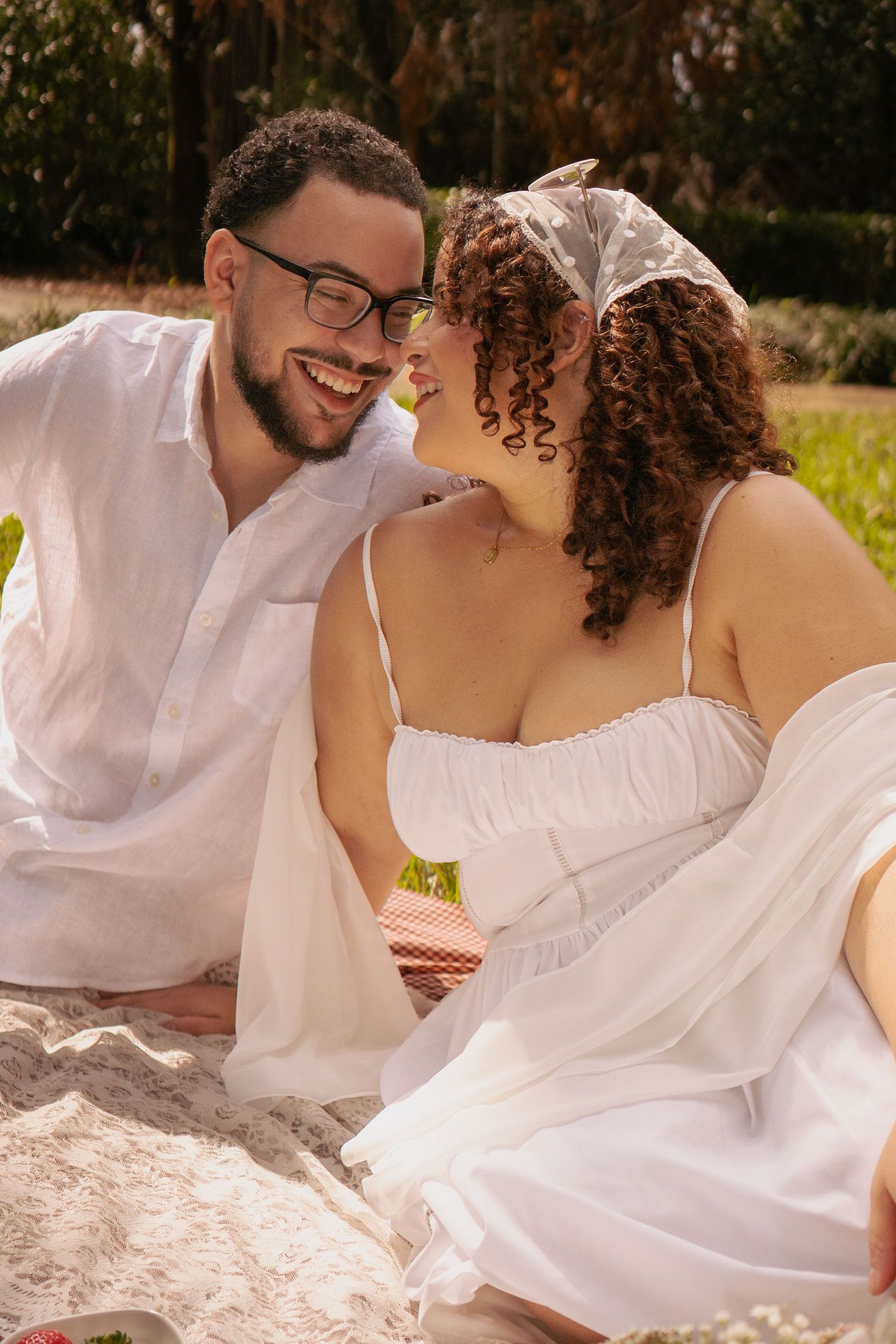 Smiling couple in white outfits sitting closely together on a blanket in a sunny outdoor setting.