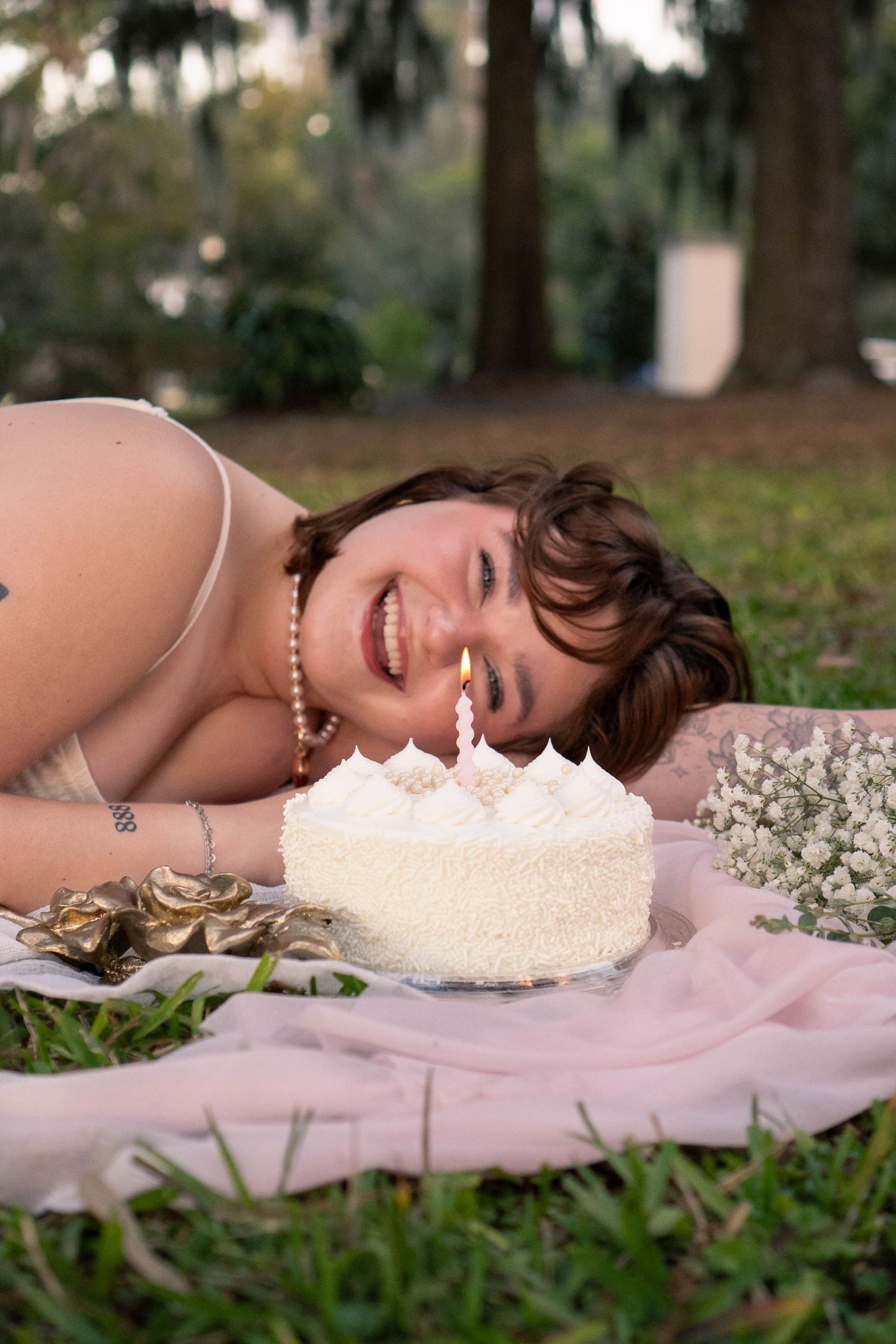 Young woman lying on grass smiling beside a white birthday cake with one candle outdoors.
