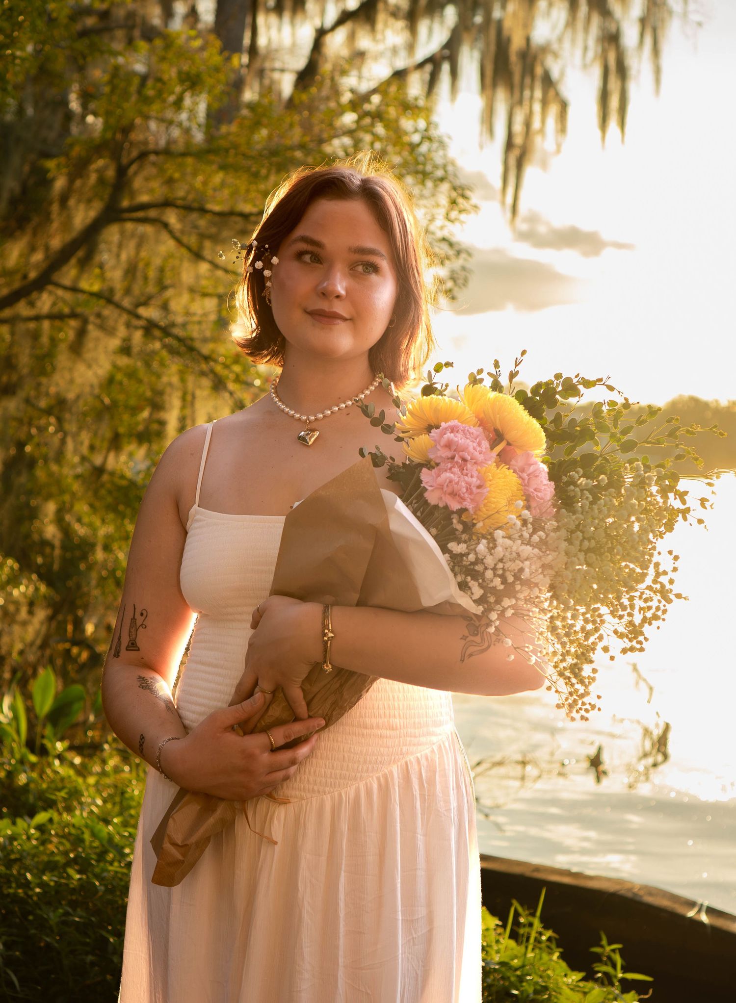 Young woman in white dress holding wrapped flower bouquet in golden sunlight near water.
