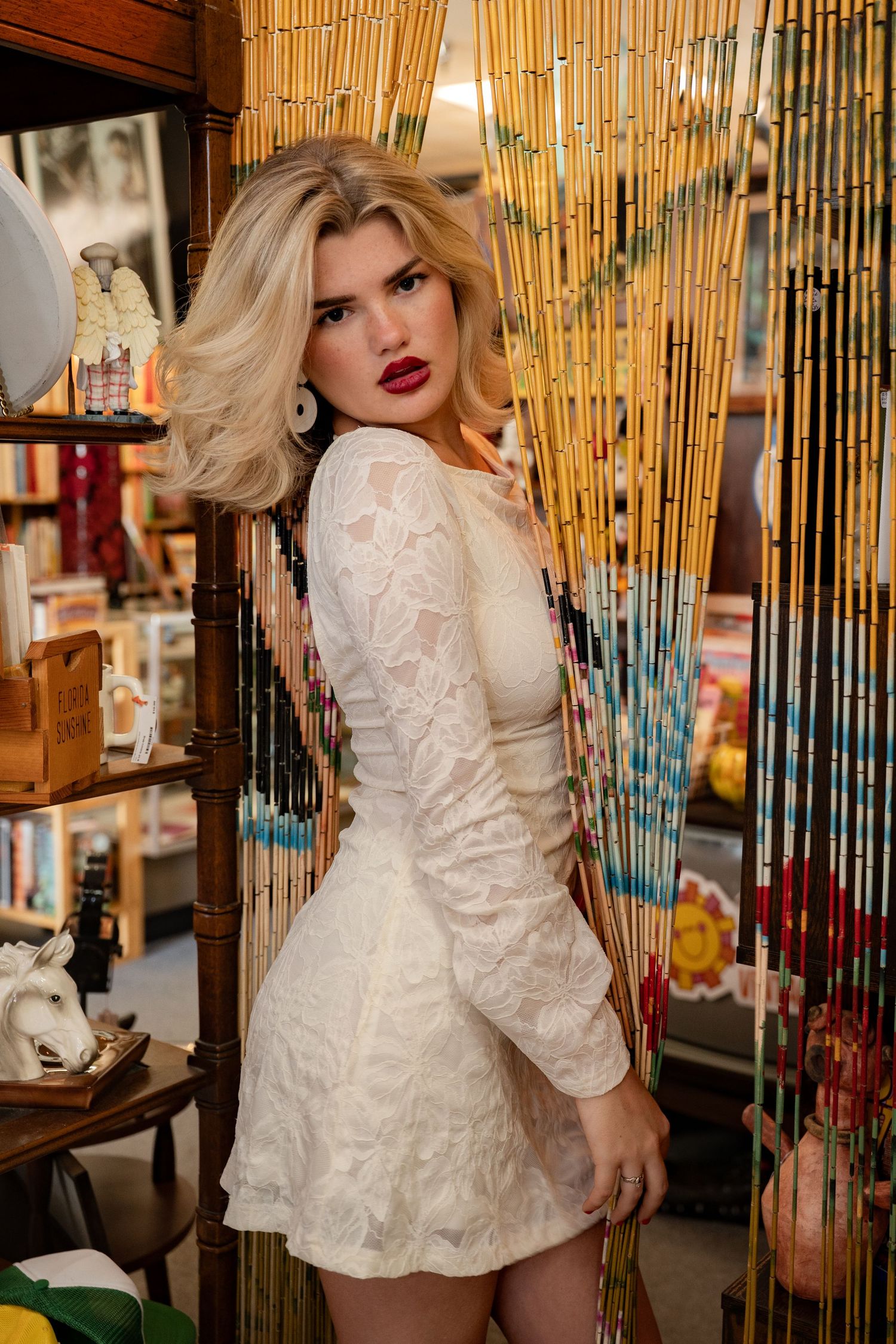 Blonde woman in white lace mini dress posing by bamboo bead curtain in eclectic vintage shop.