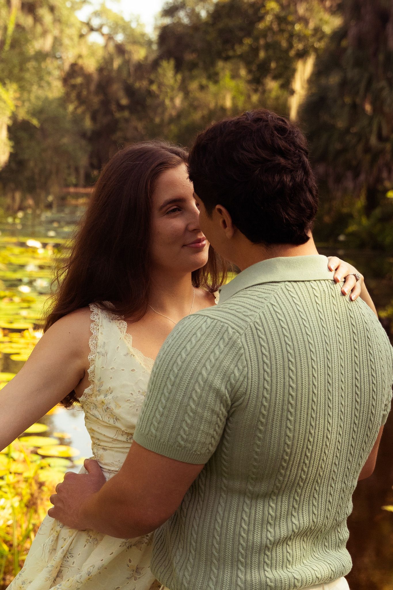 Couple embracing near a sunlit pond surrounded by lush greenery, woman in white dress smiling warmly.