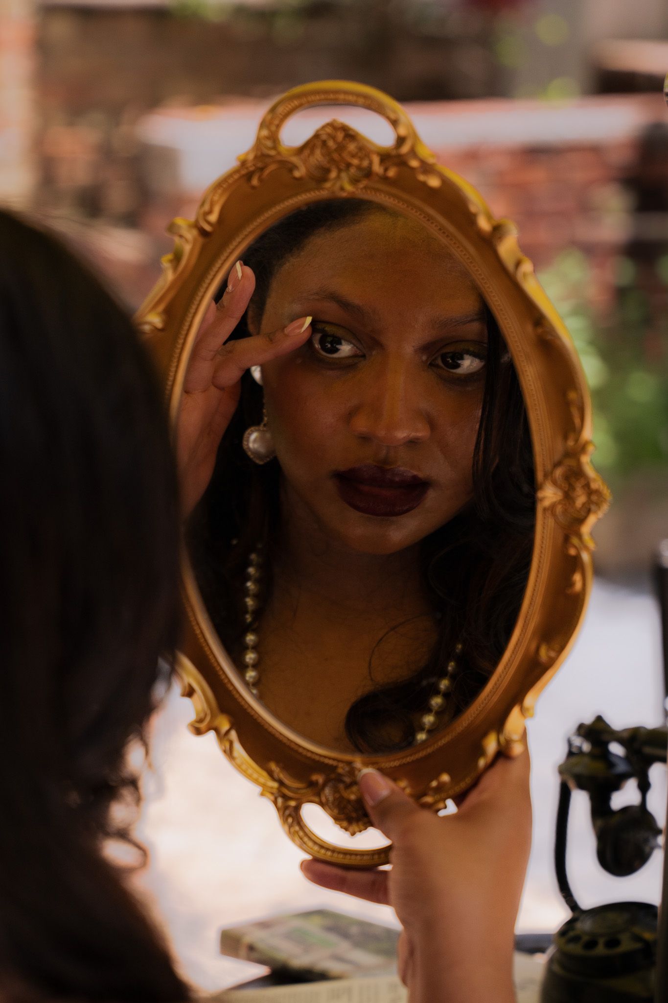 Woman with dark lipstick and pearl necklace gazing into ornate gold oval mirror in moody setting.
