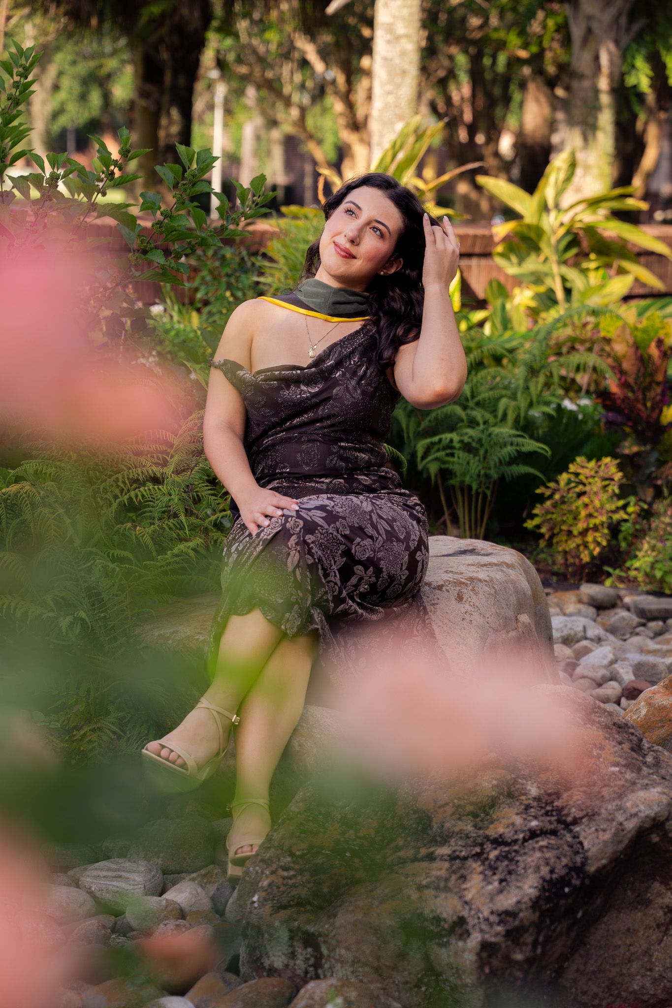 Woman in dark velvet dress sits on rocks surrounded by lush tropical garden greenery.