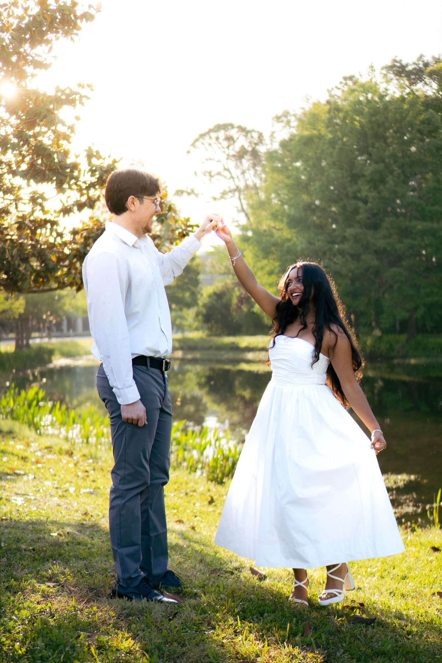 Couple dancing outdoors at golden hour near a pond, woman in white dress being twirled.