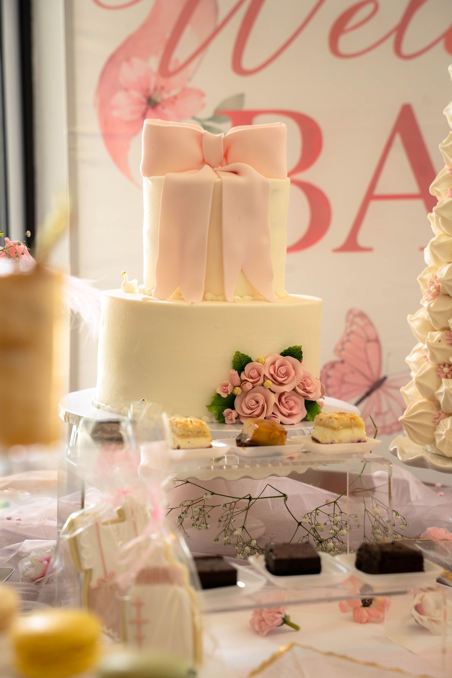 Elegant two-tier white baby shower cake with pink bow and rose decorations on a dessert table.