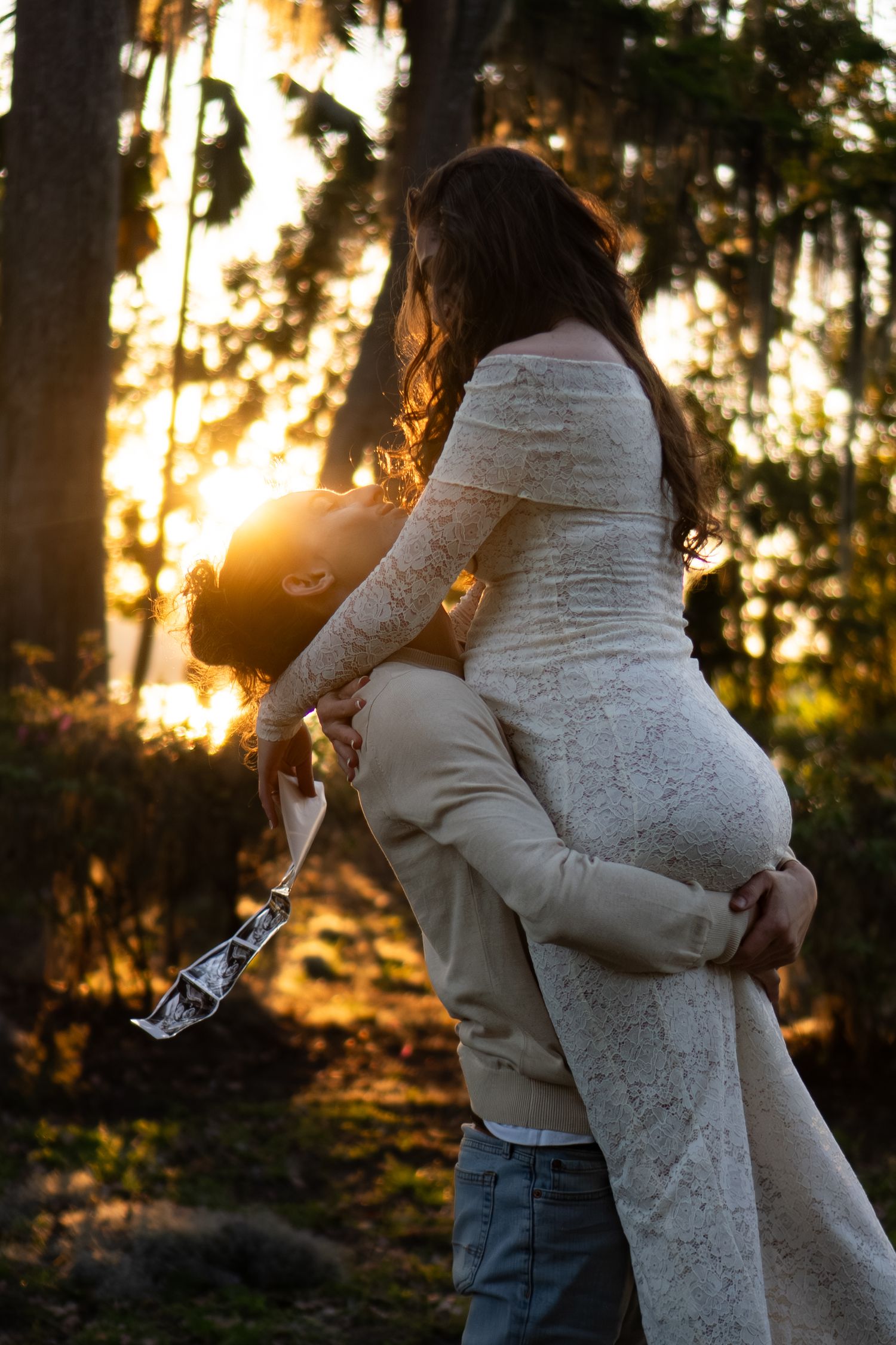 Man lifting pregnant woman in white lace dress in a sunlit forest during a glowing golden sunset.