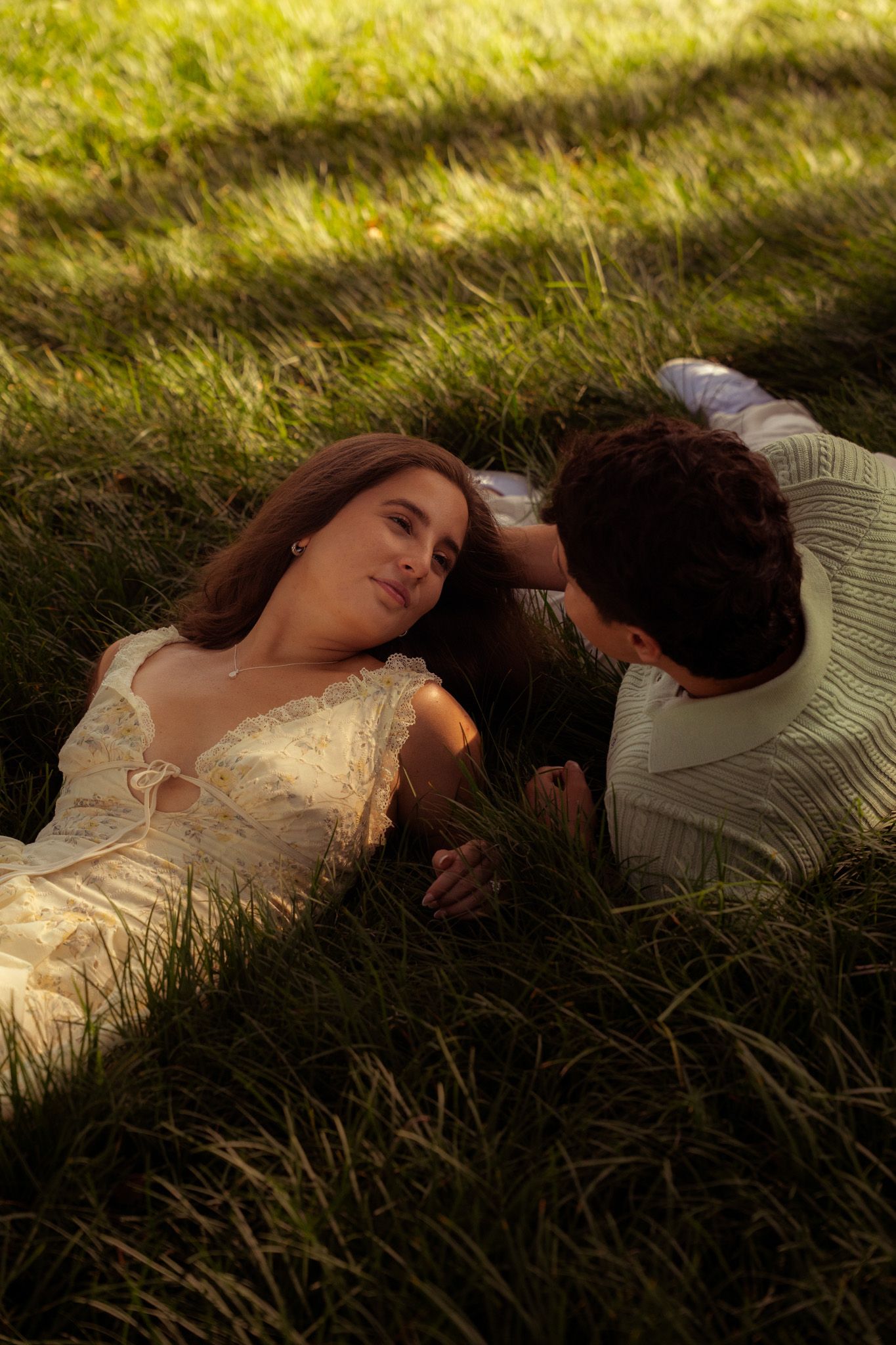 Young couple lying in lush green grass holding hands, gazing at each other in warm sunlight.