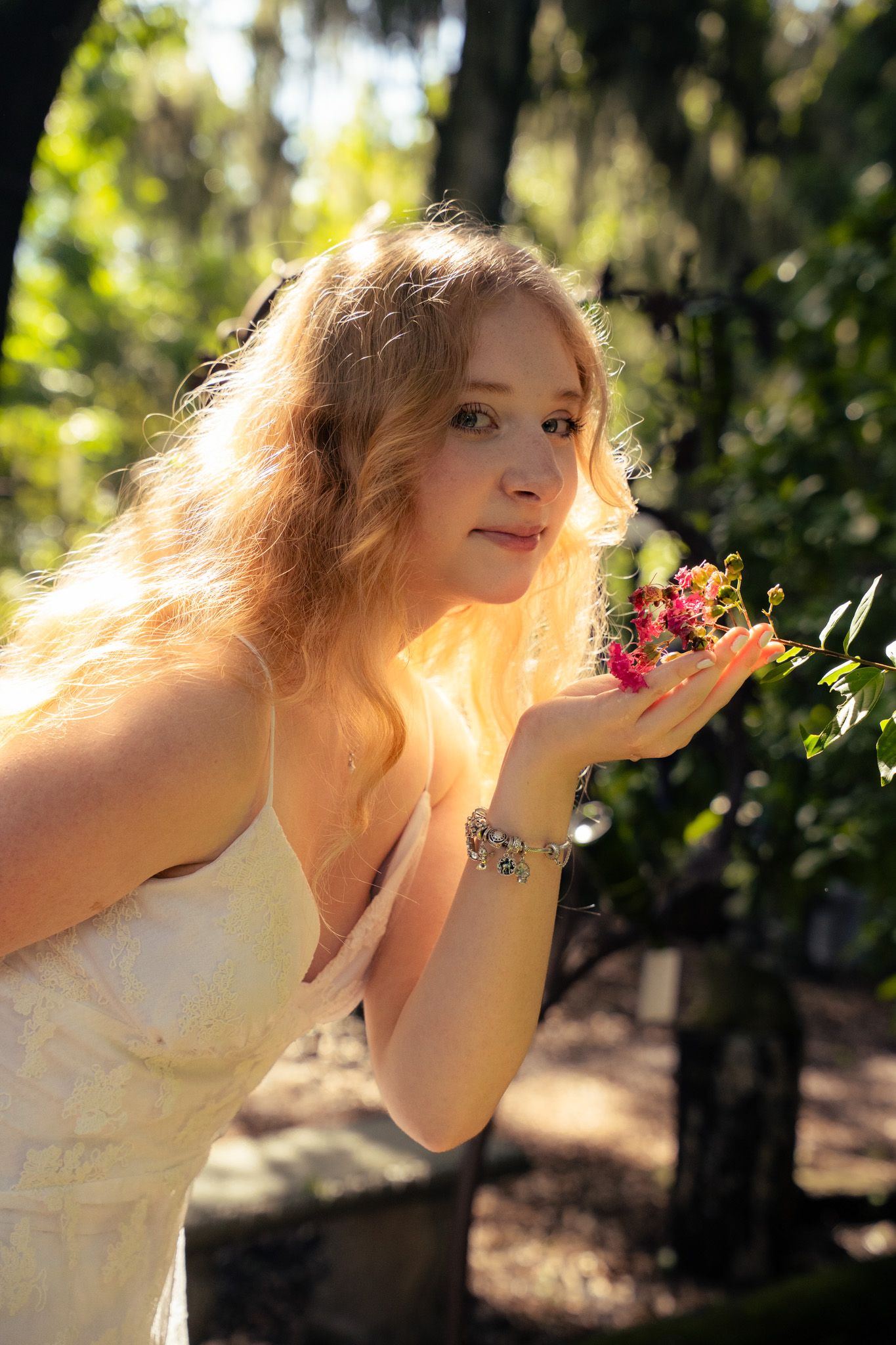 Beautiful blonde woman in white dress holding pink flowers in a sunlit green garden setting.