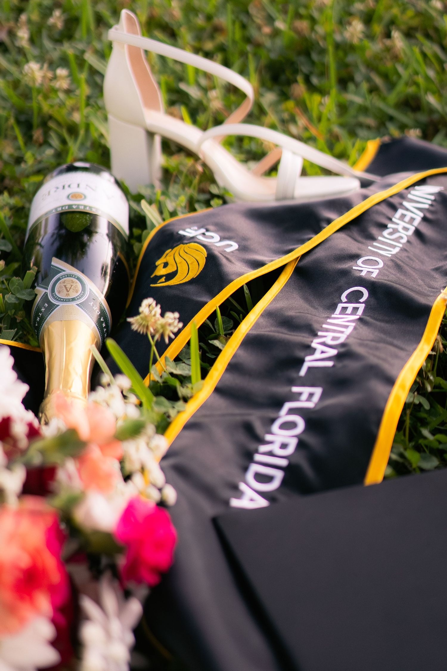 UCF graduation stole, champagne bottle, white heels, and flowers arranged on green grass.