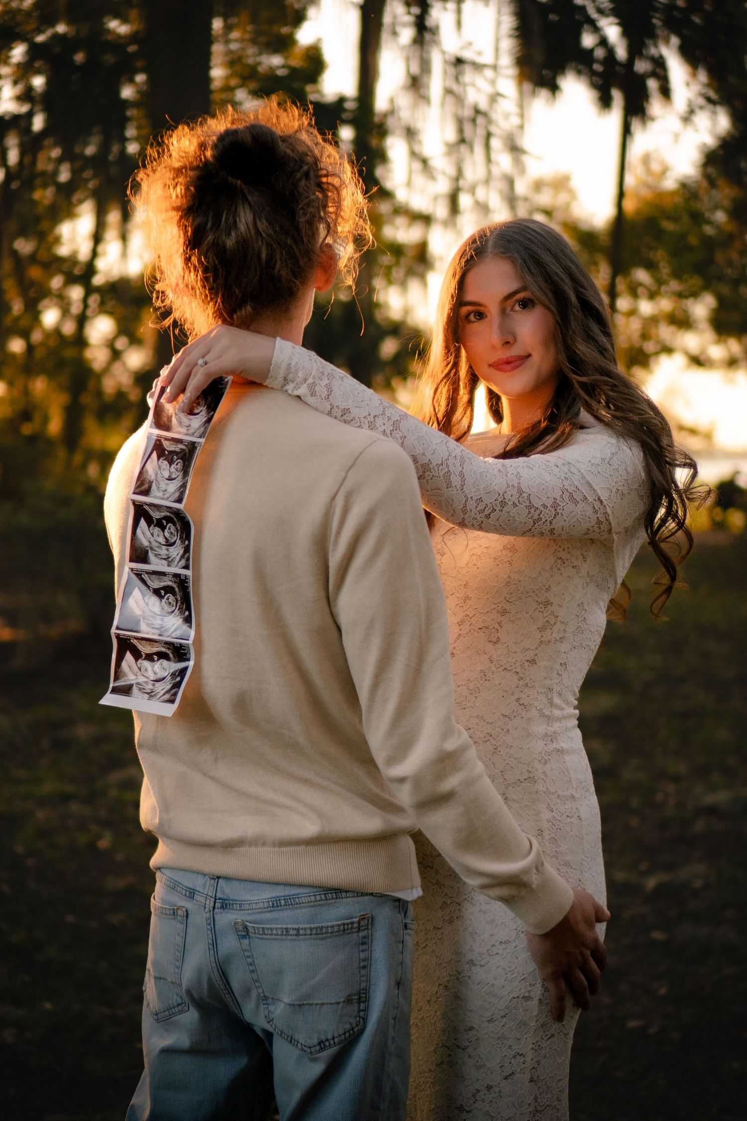 Pregnant woman in white lace dress embracing partner who holds ultrasound strip in golden forest light.