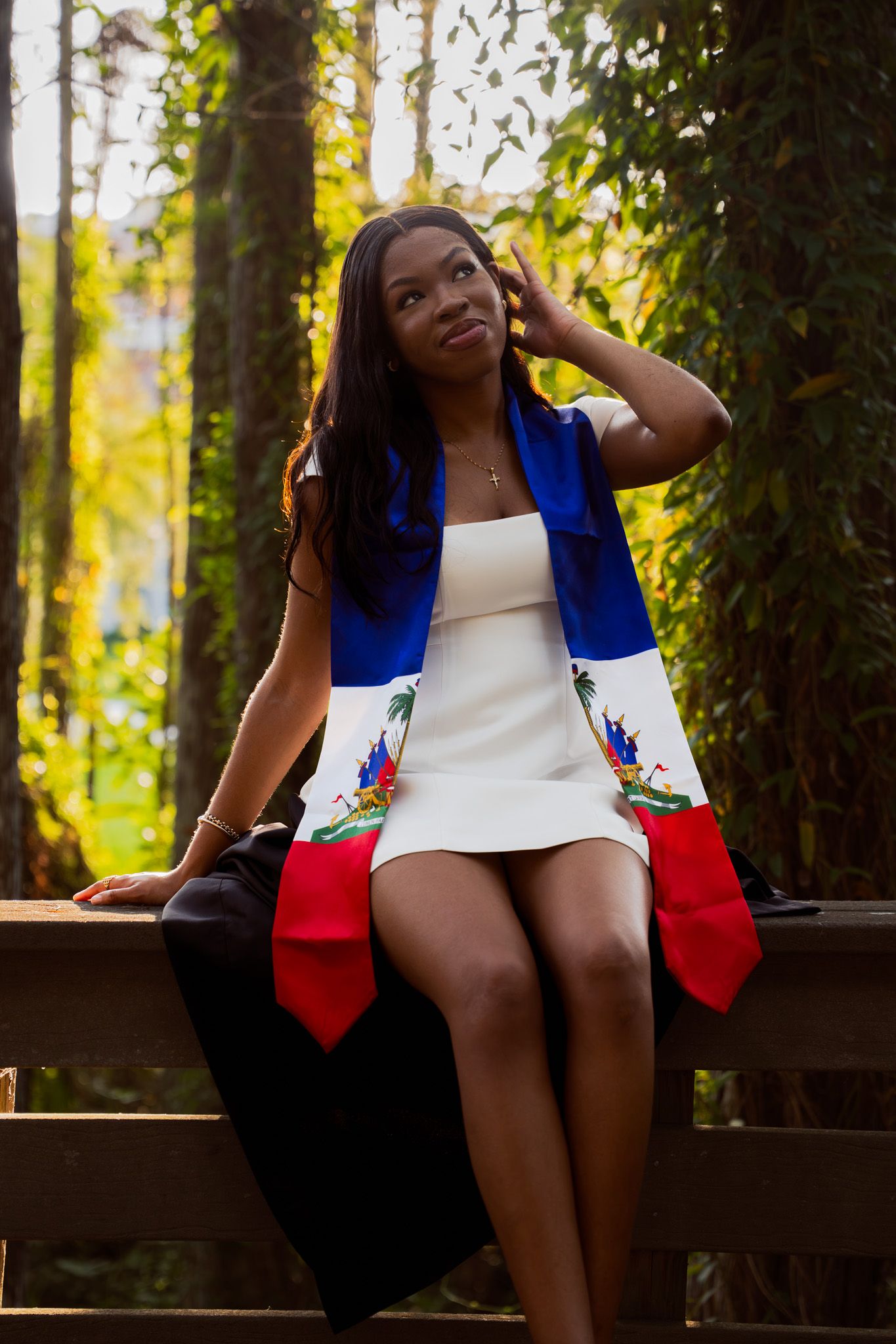Young woman in white dress with Haitian flag draped over shoulders sits on fence in forest setting.