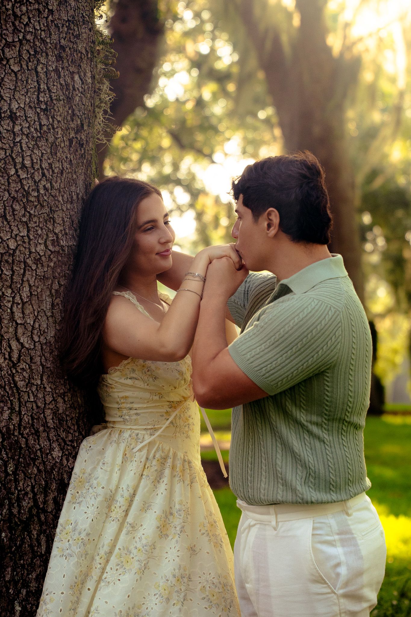 Couple sharing a romantic moment by a tree, man kissing woman's hand at sunset.