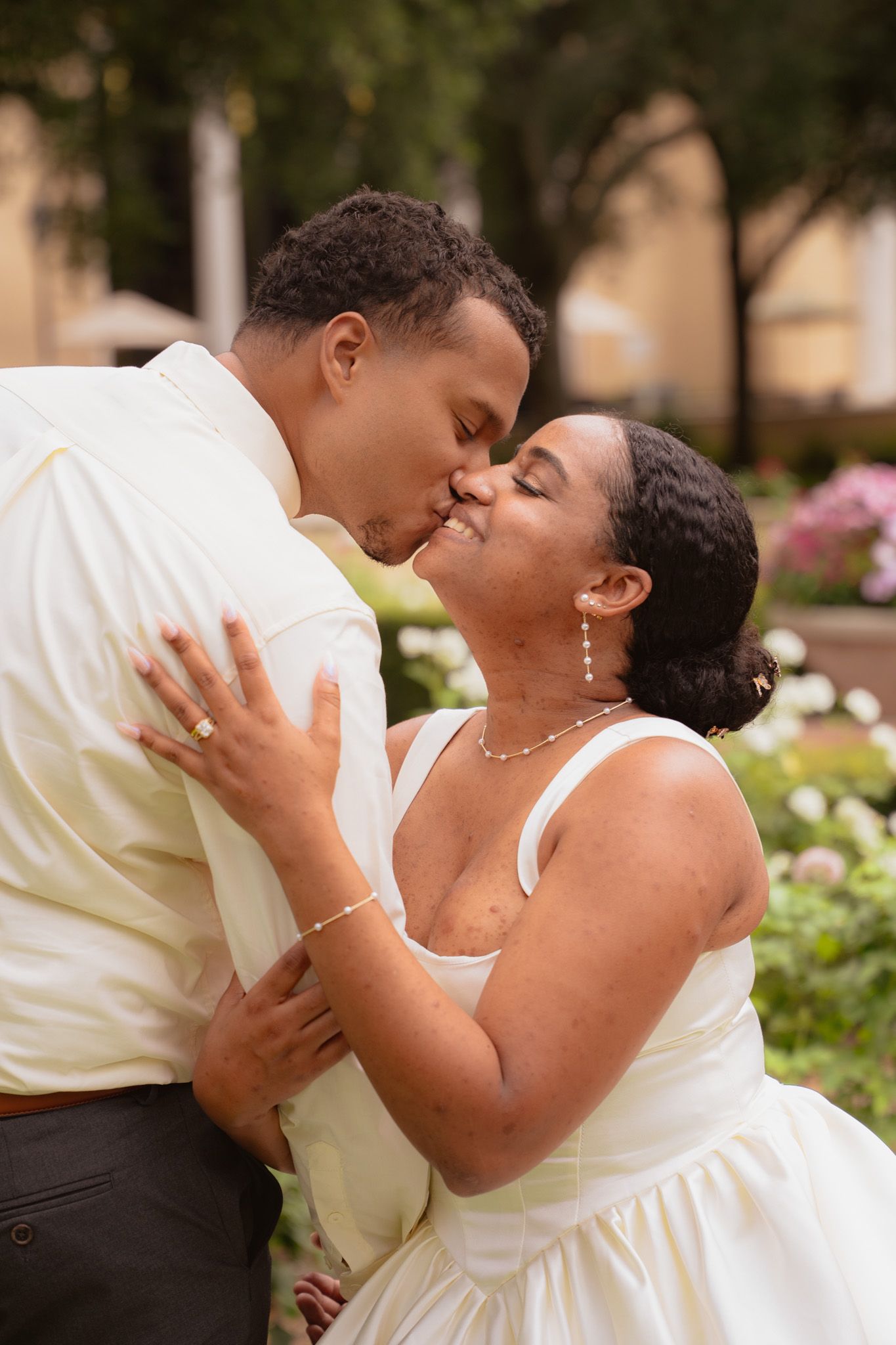 Newlywed couple shares a romantic kiss outdoors surrounded by lush garden greenery.