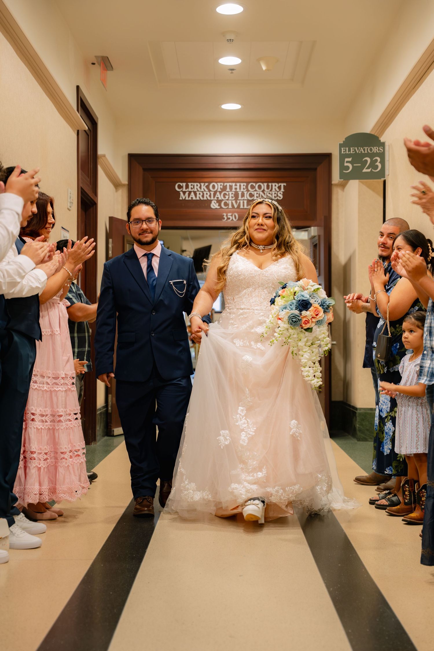 Newlyweds walk hand-in-hand through courthouse hallway as guests applaud after civil marriage ceremony.