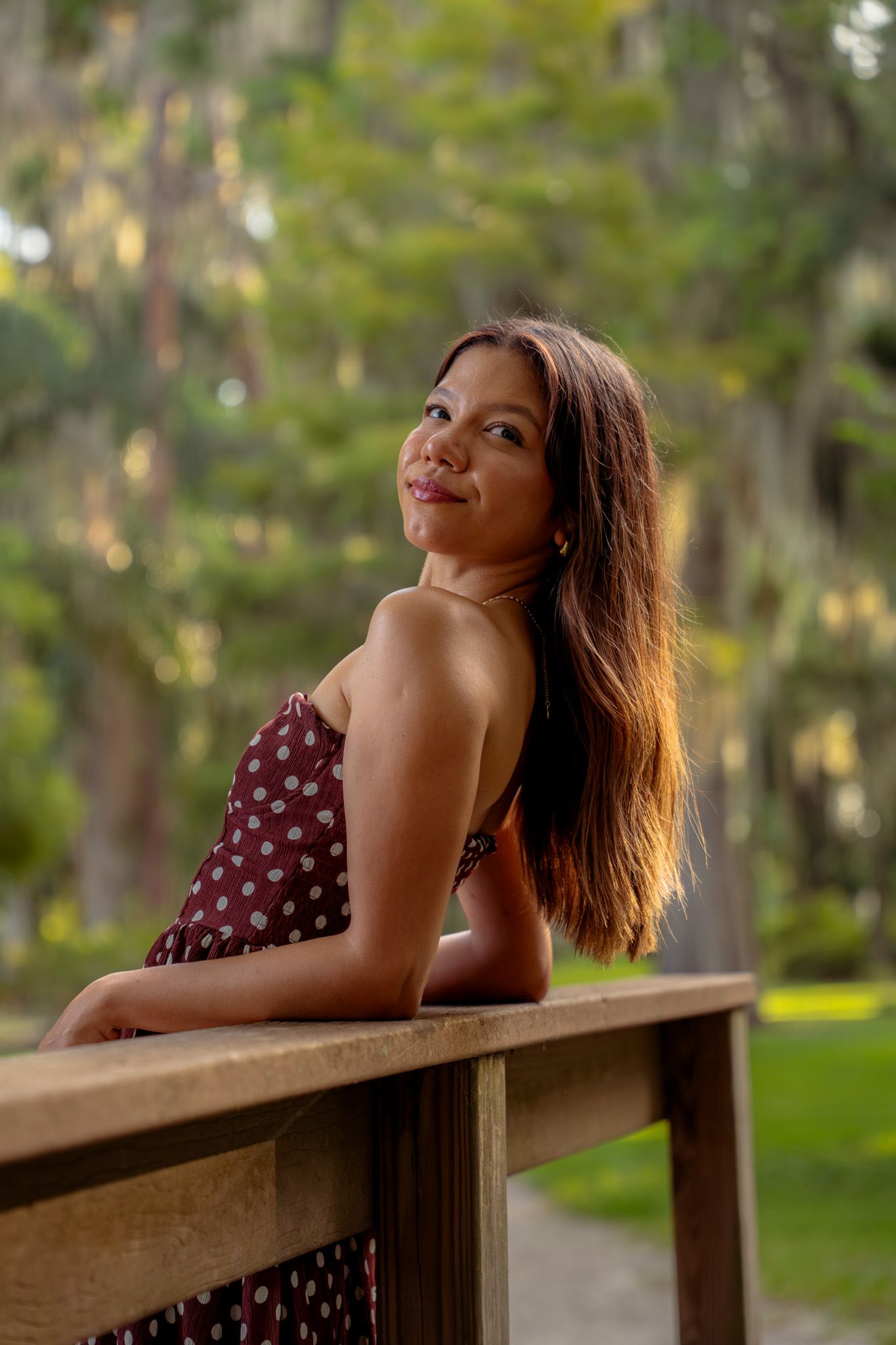 Smiling young woman in polka dot sundress leaning on wooden railing in sunlit park with tall trees.
