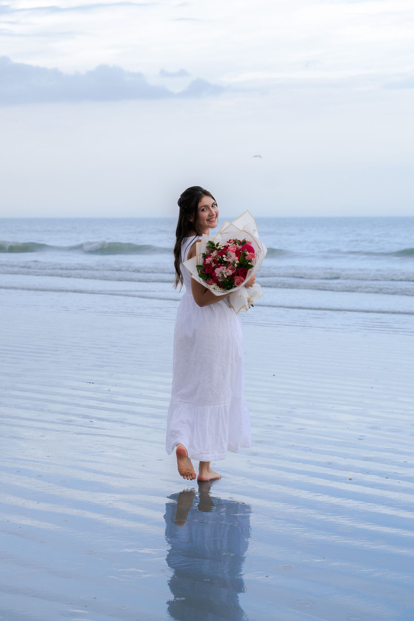 Woman in white dress standing on wet beach holding a large bouquet of pink flowers.