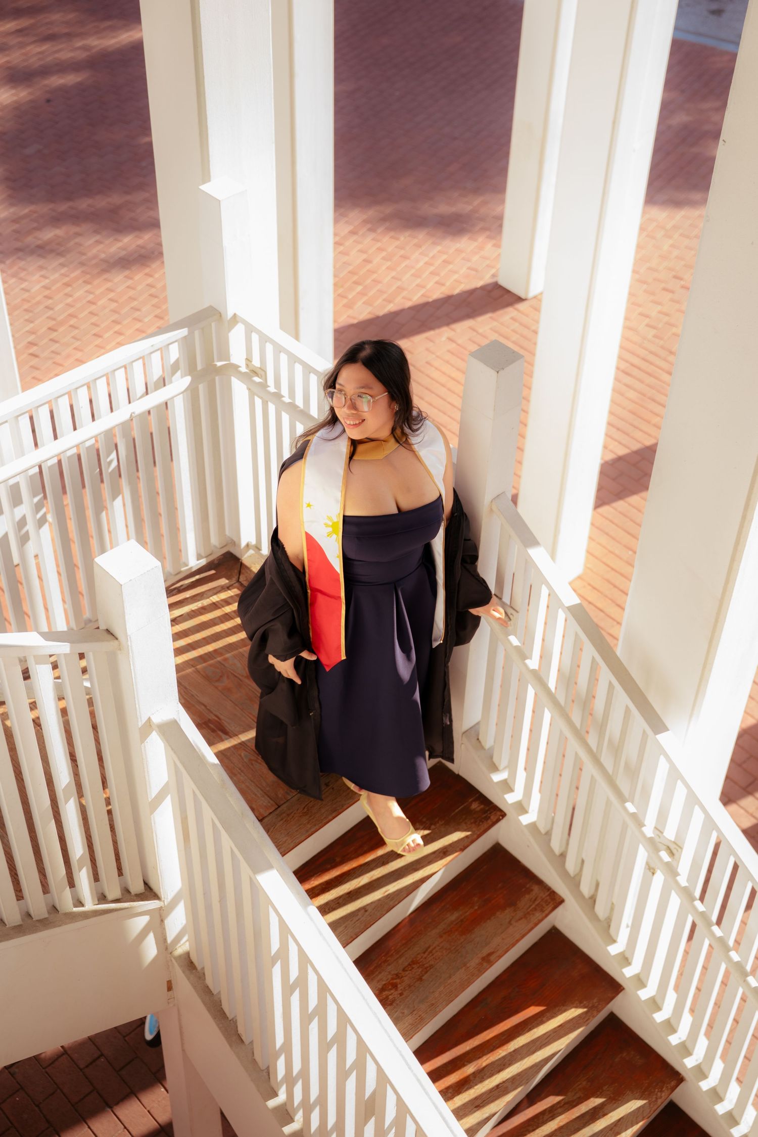 Graduate in navy dress and black gown with colorful hood poses on white spiral staircase.