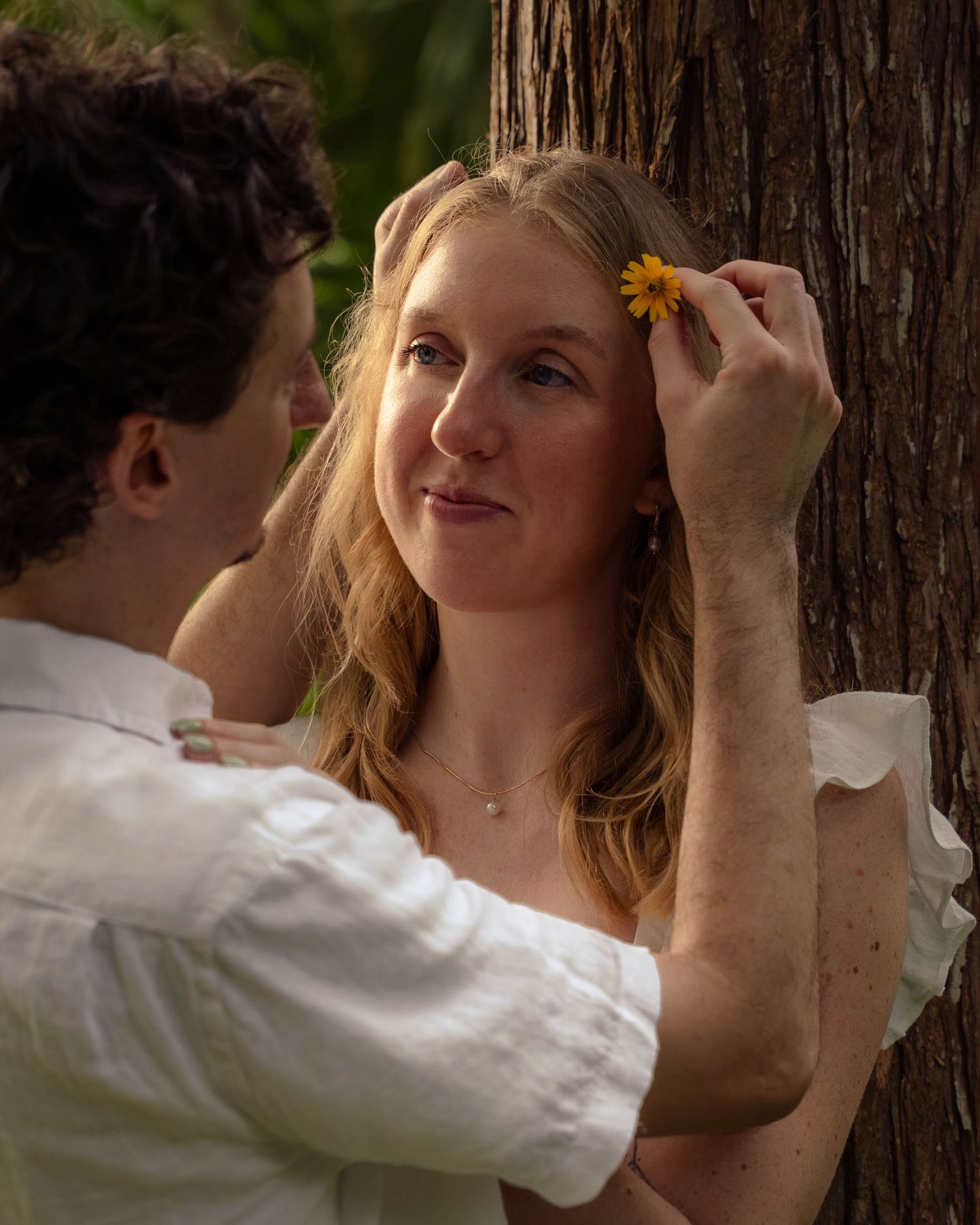 Man placing a yellow flower in a smiling woman's hair near a tree in a park.